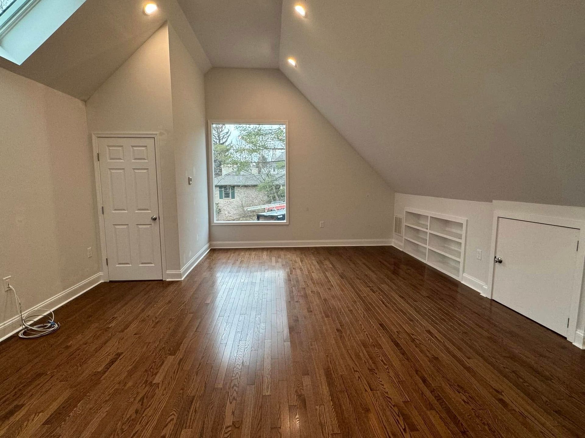 Empty attic room with hardwood floor, slanted ceiling, window, and door.