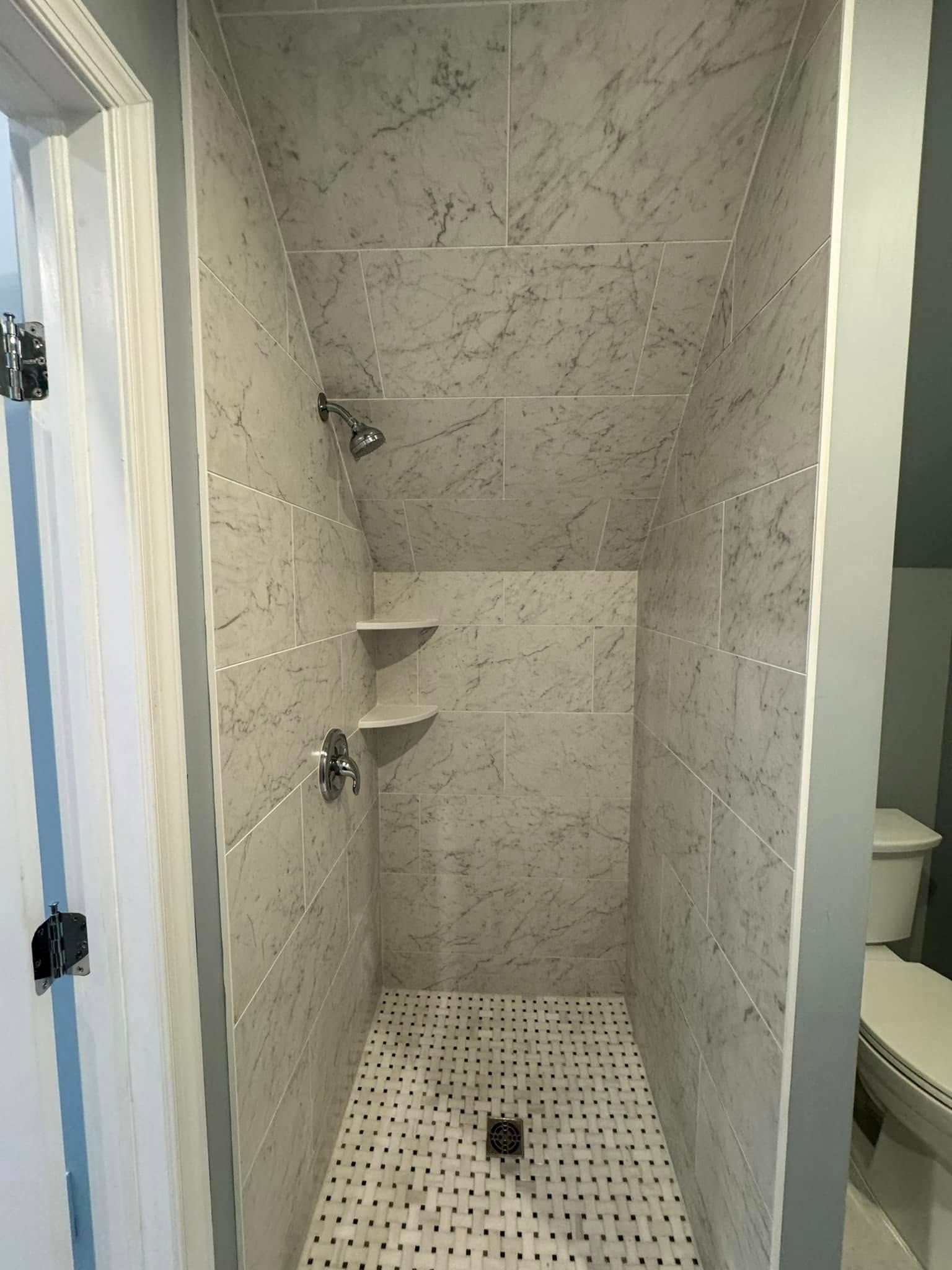 Narrow shower with gray tile, two shelves, two showerheads, and a toilet to the right.