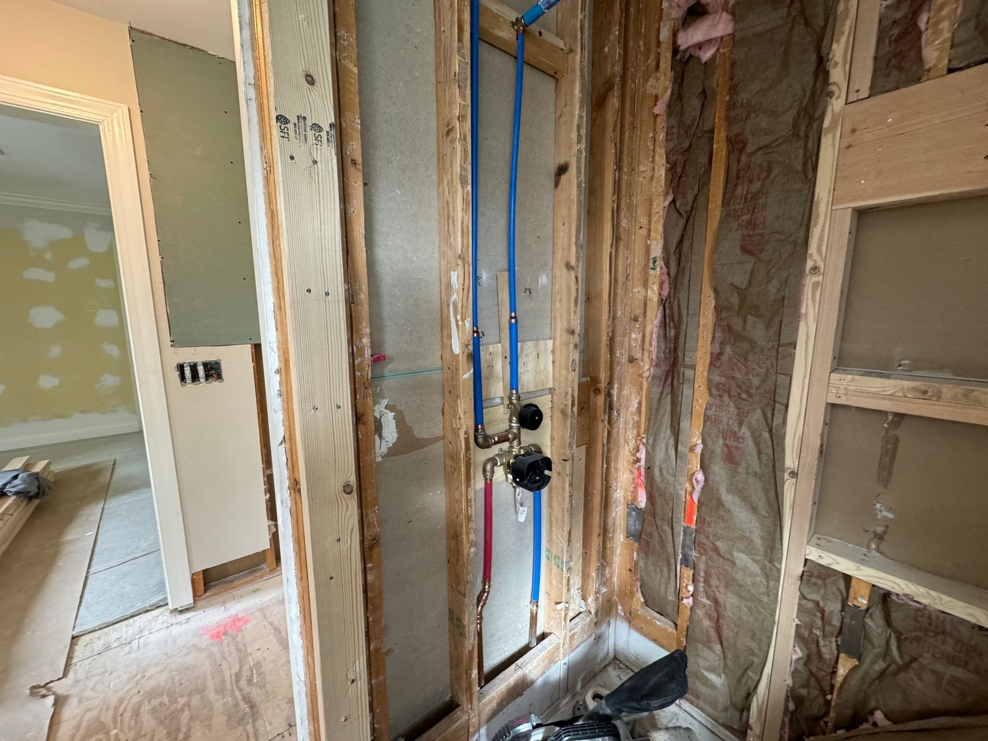 Bathroom under construction, with exposed framing, blue pipes, and shower fixtures installed.
