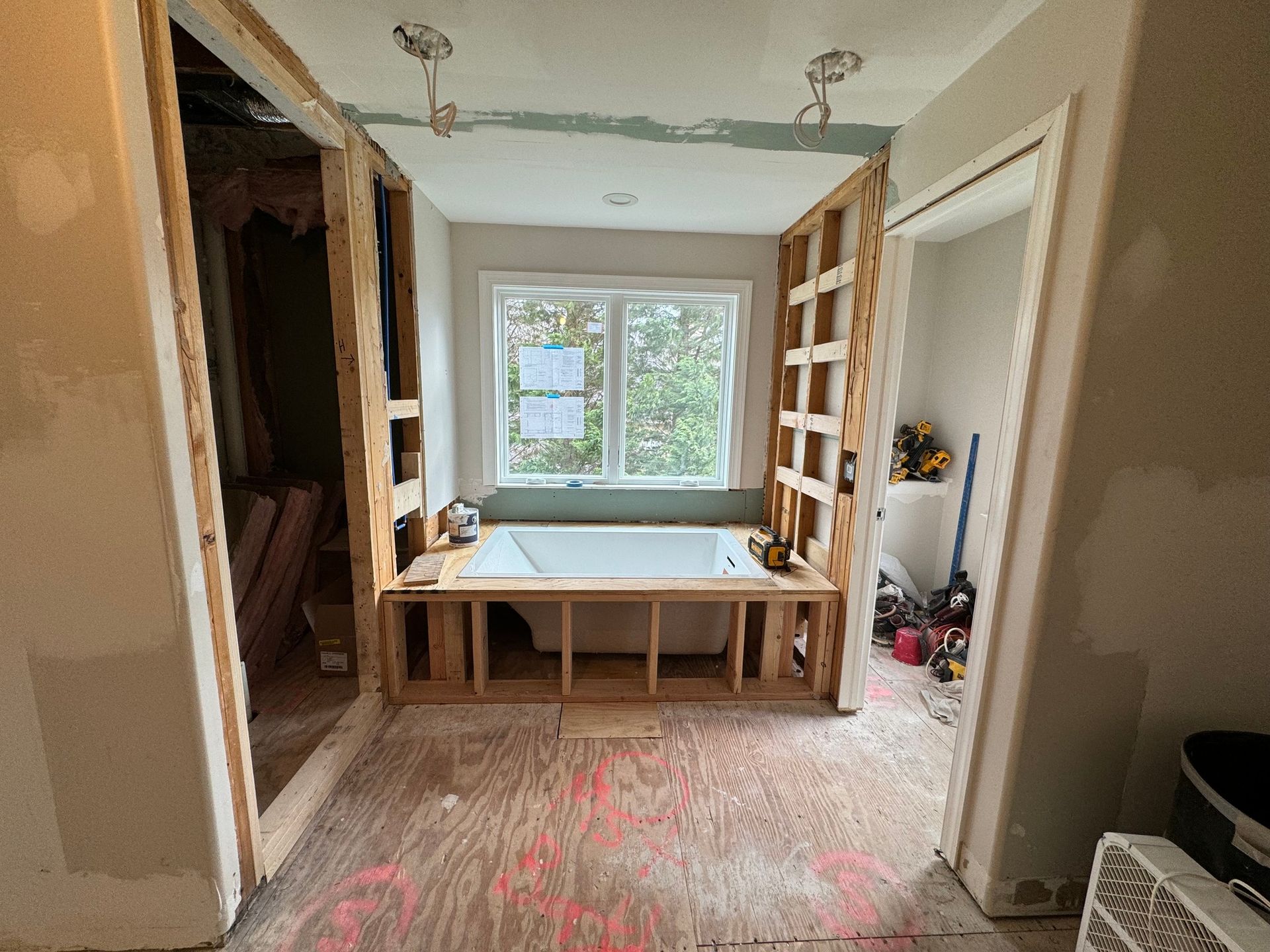 Bathroom undergoing renovation; wooden frames surround a bathtub under a window; exposed studs and construction materials.