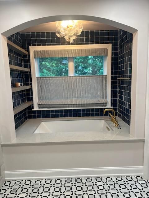 Bathroom with a built-in tub, blue tile, shelves, window, and gold fixtures. Black and white patterned floor.