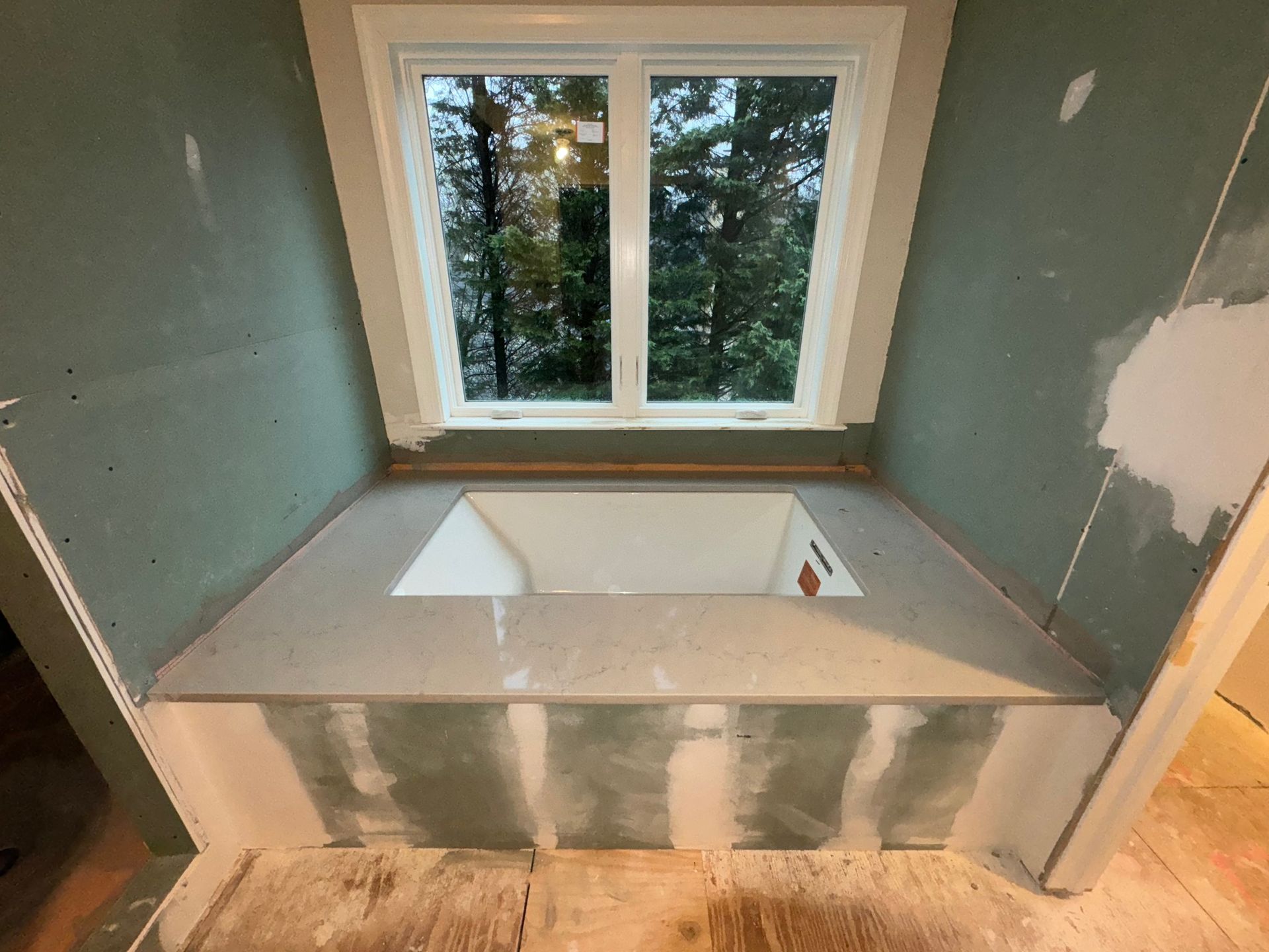 Bathroom under construction: a built-in tub with a window above. Gray walls, unfinished drywall, and a view of trees.