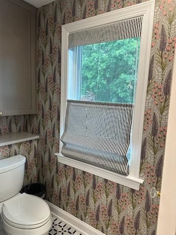 Bathroom with floral wallpaper, a window with striped shade, toilet, and cabinet.