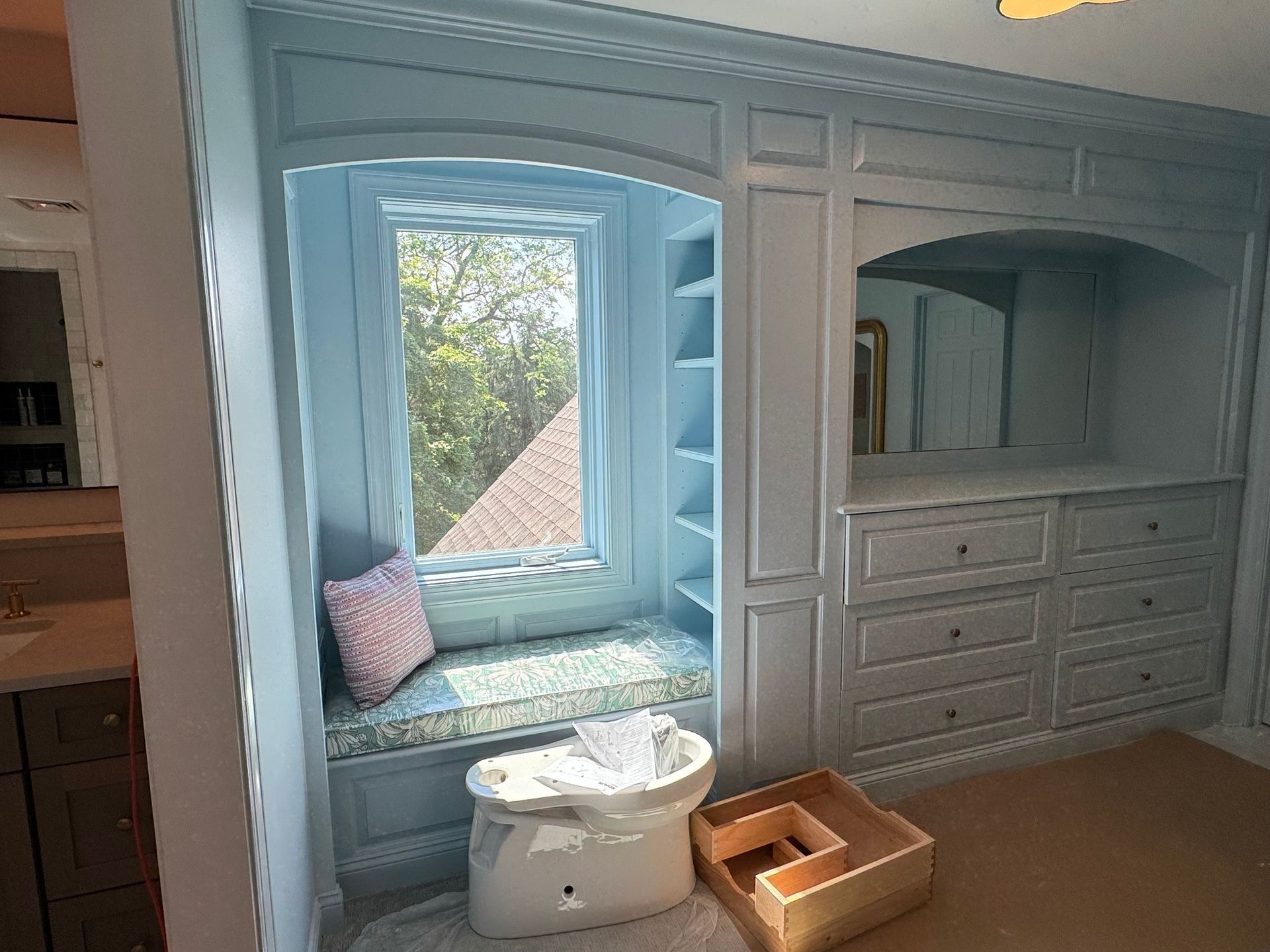 Built-in light blue window seat with shelves, drawers, and arched mirror. Green foliage visible through the window.