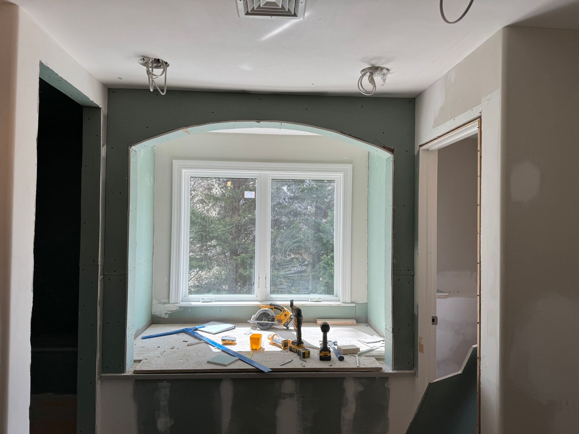 Interior shot of a window with an arched top, surrounded by unfinished drywall and construction tools on the sill.