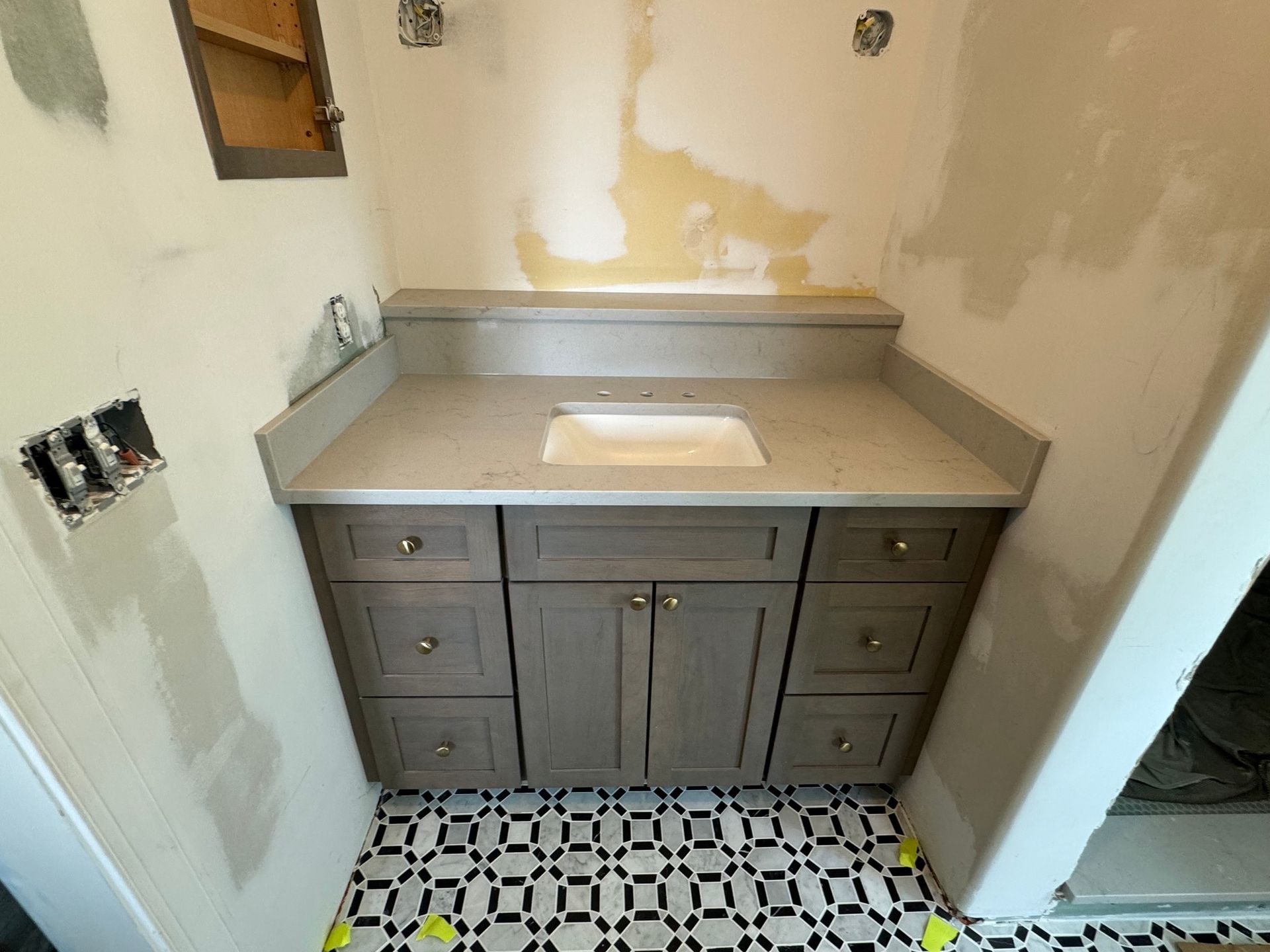 Bathroom vanity with gray cabinets, light countertop, and black and white patterned tile. Walls are unfinished.