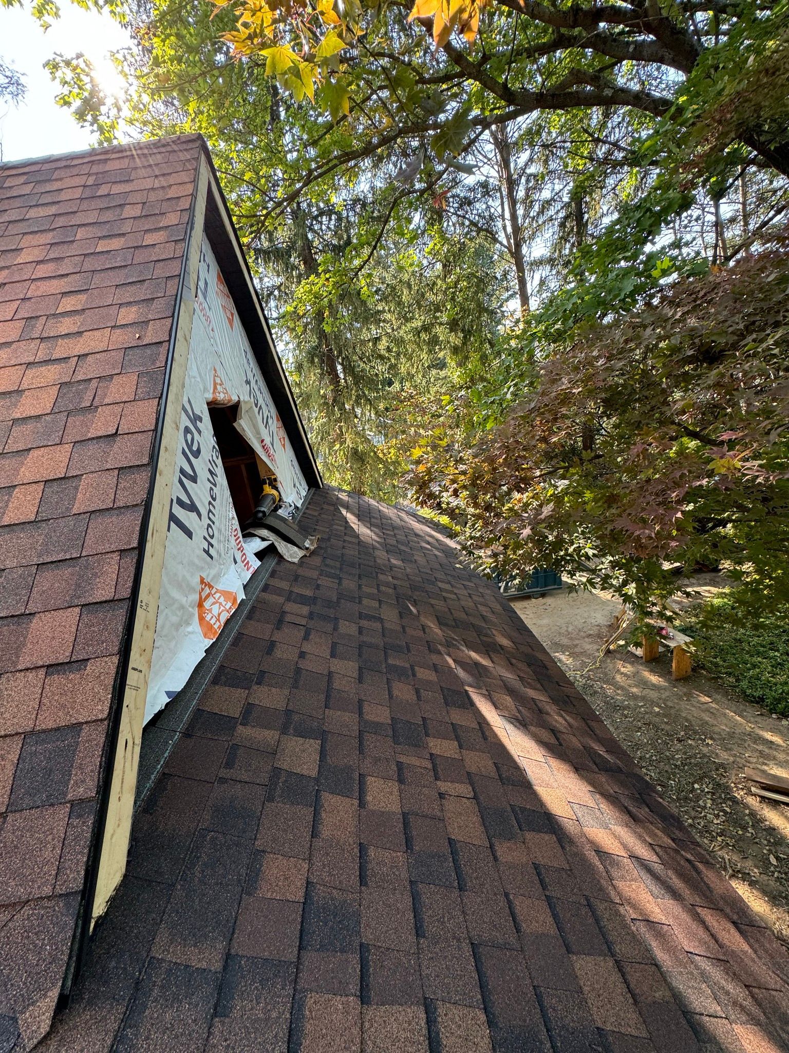 Roof with brown shingles, unfinished gable end, Tyvek wrap visible, trees in background.