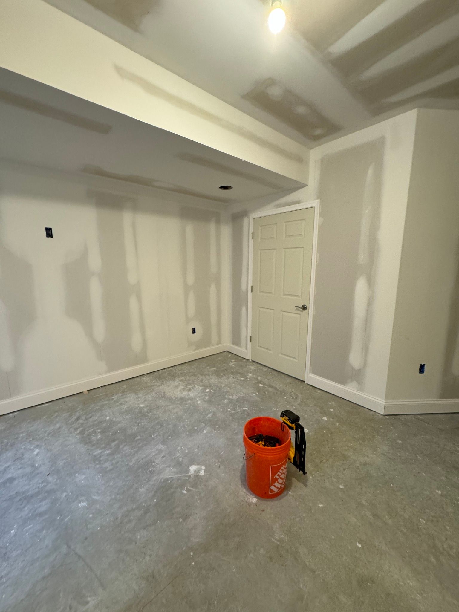 Room under construction, drywall installed, concrete floor, orange bucket, and door.