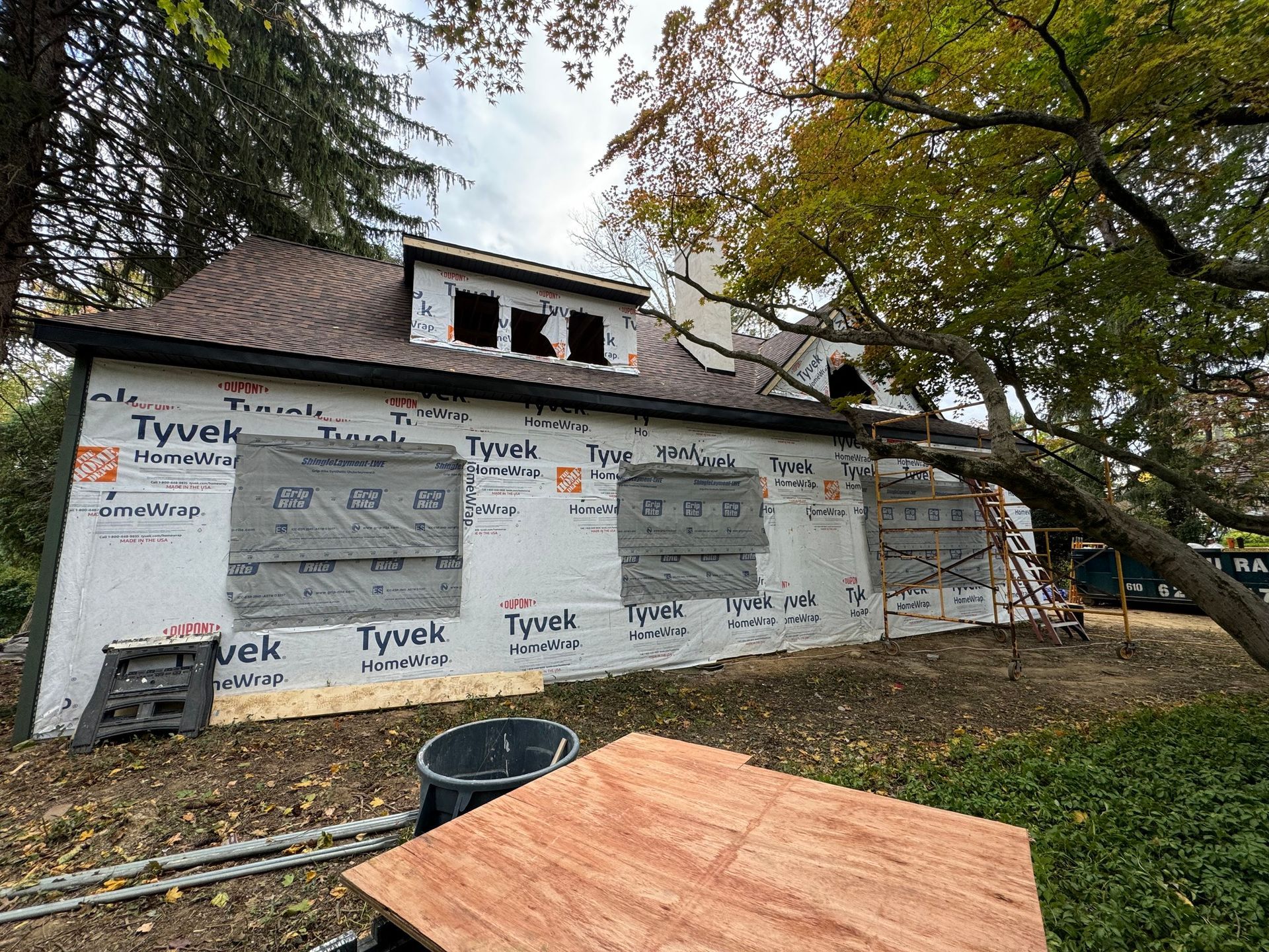 House under construction, Tyvek wrap on exterior, brown roof, windows, and surrounding trees.