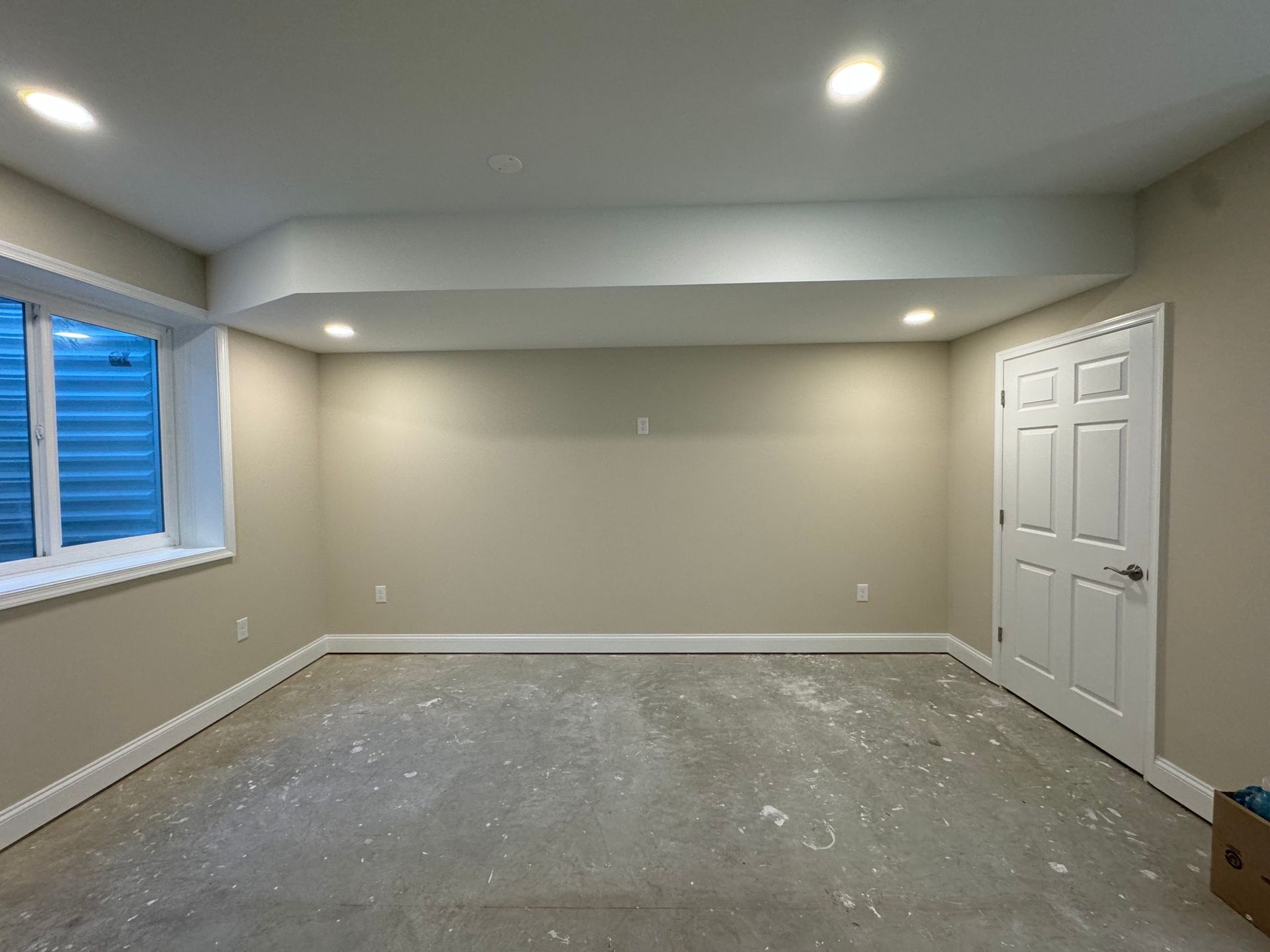 Empty room with beige walls, white trim, recessed lighting, and a white door. The concrete floor shows splatters.