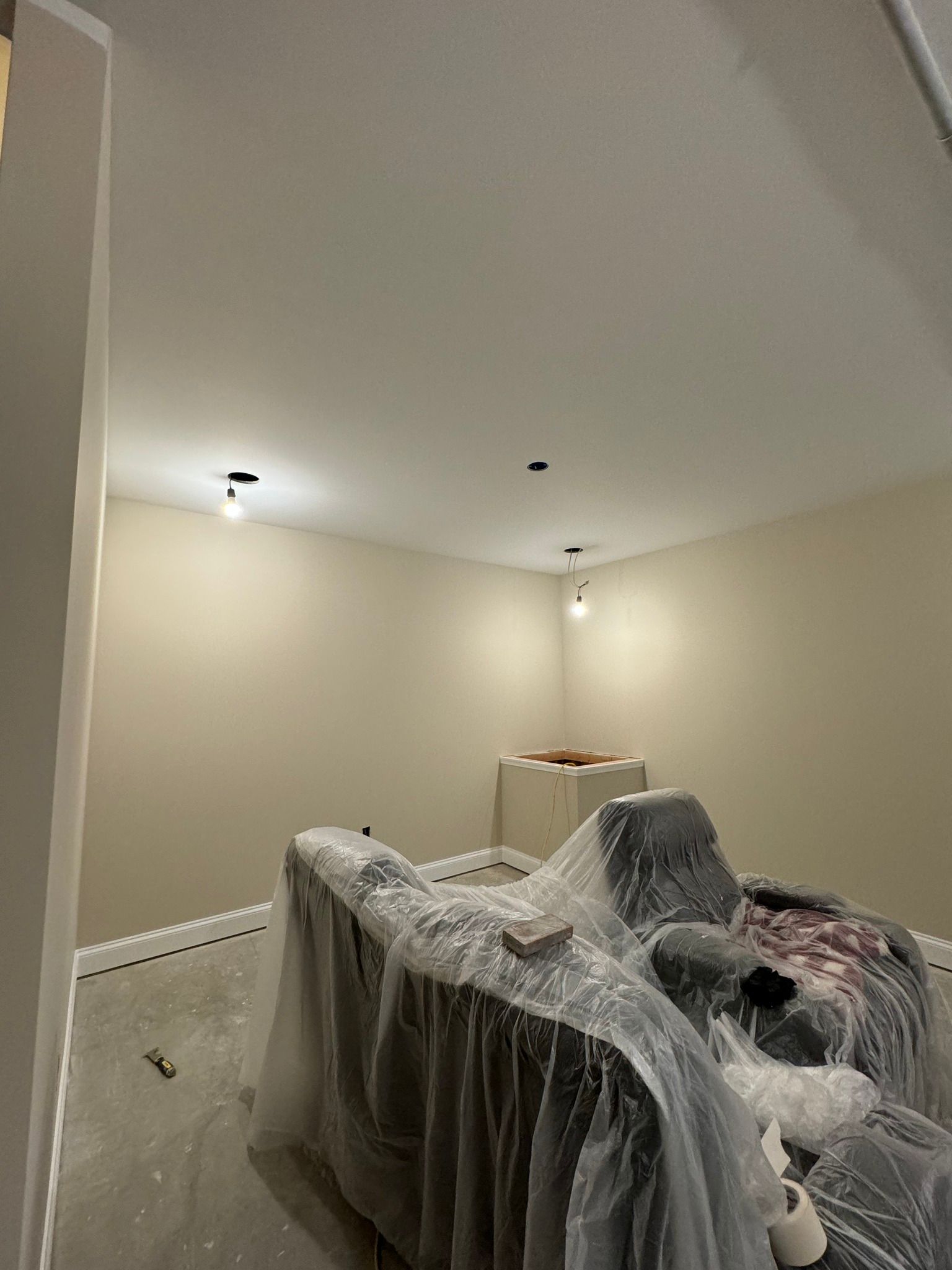 Interior room with beige walls, two lights, covered furniture, unfinished floor.