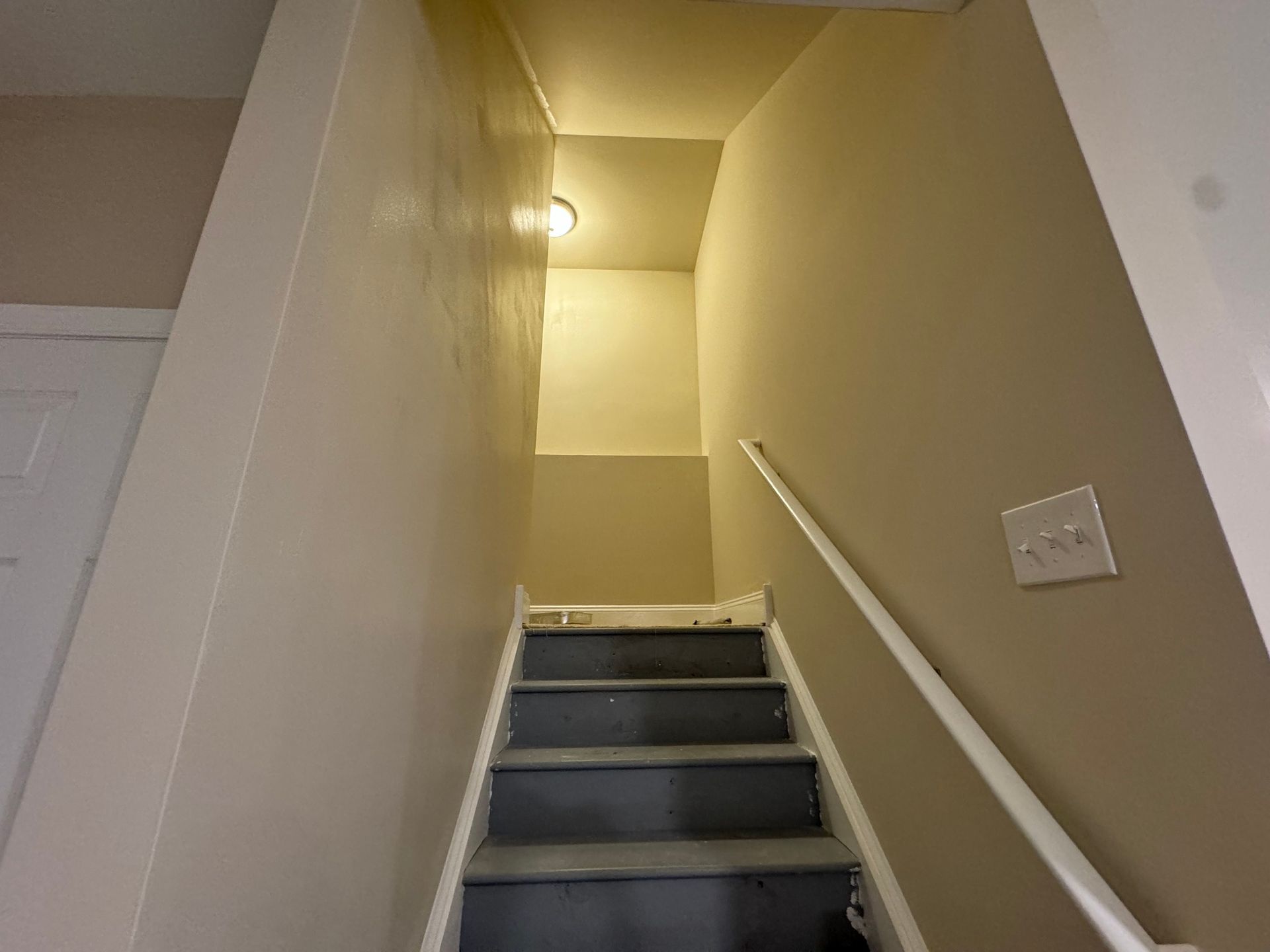 Staircase with gray steps and white handrail leading up to a small, lit landing; beige walls.