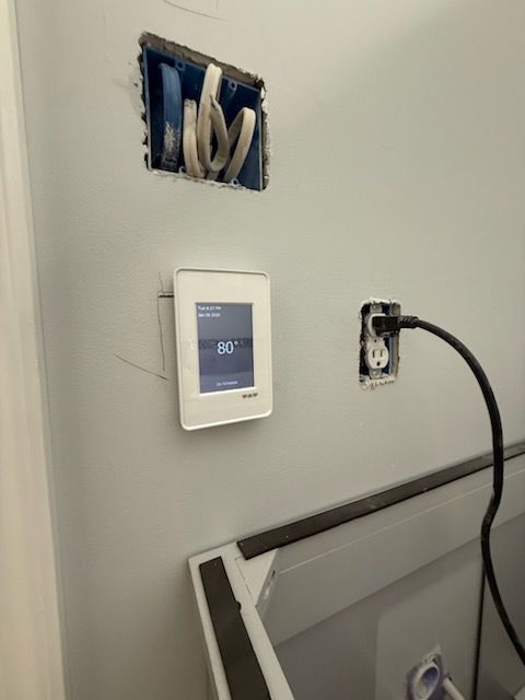 White smart thermostat on wall, next to open electrical box and power outlet. Gray wall, indoor setting.