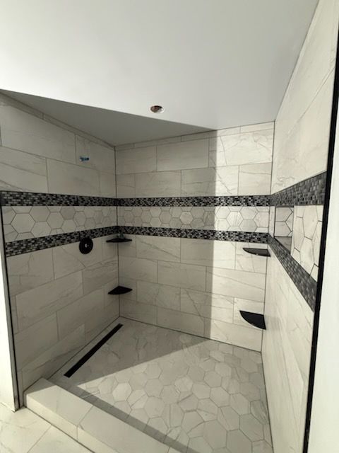Tiled shower stall with white walls, black accent tile band, three black corner shelves, and a linear drain.