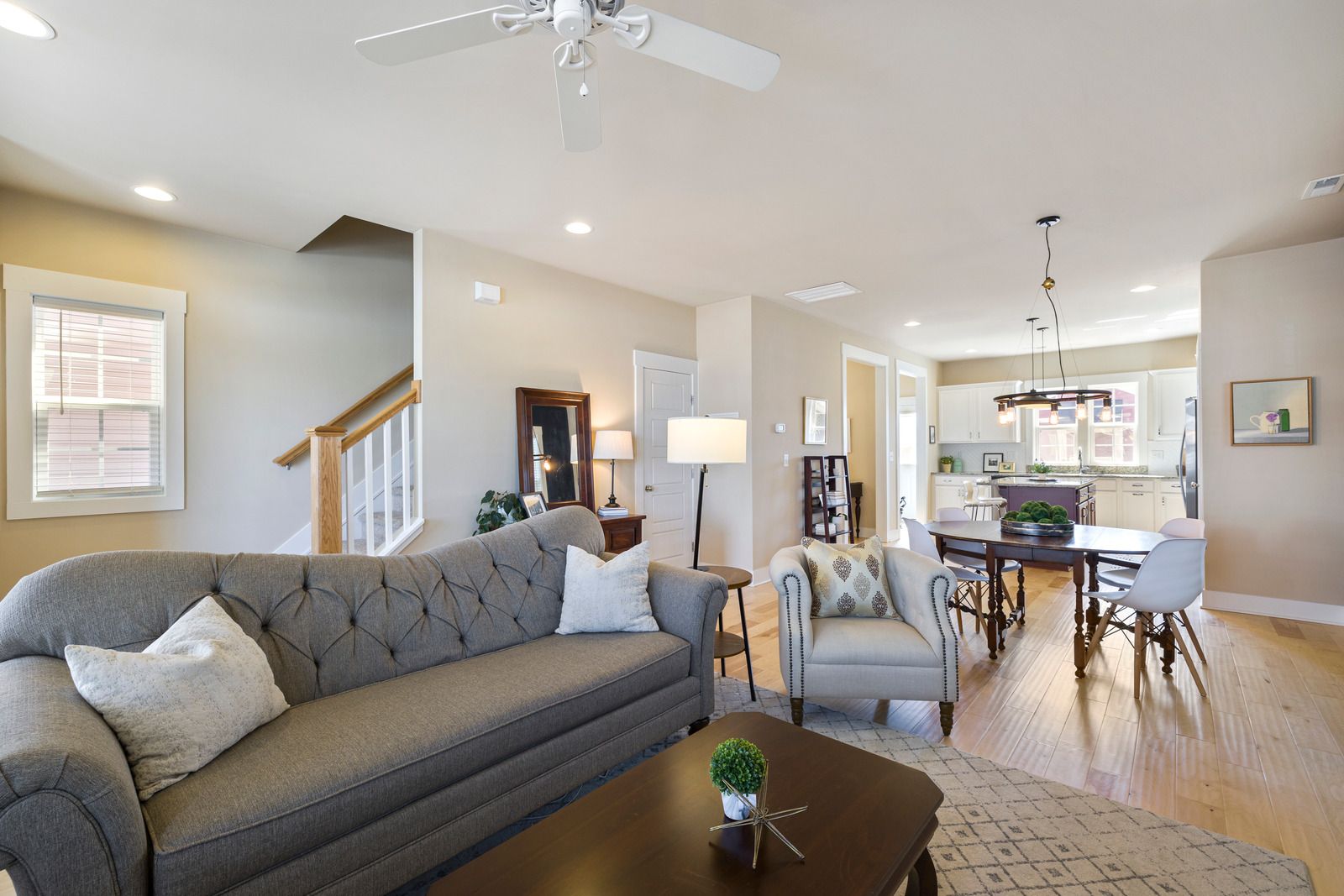 Living room with a gray tufted sofa, armchair, table, rug, and view of the dining area and kitchen.