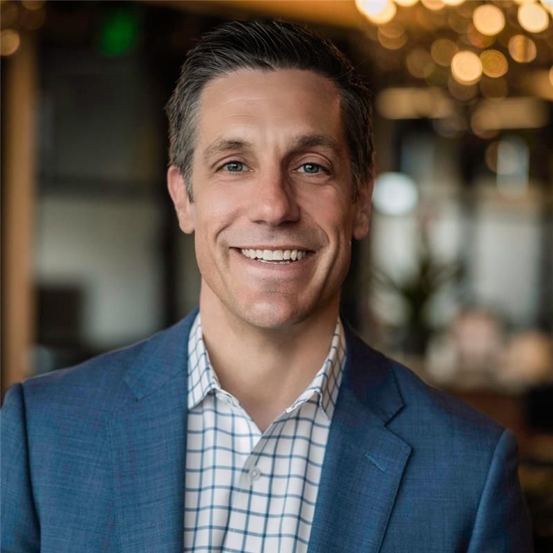 Man smiling, wearing a blue blazer and checkered shirt, indoors, soft lighting.