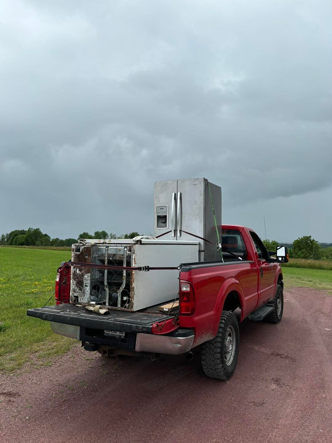 A red truck with a refrigerator and freezer in the back is parked on a dirt road.