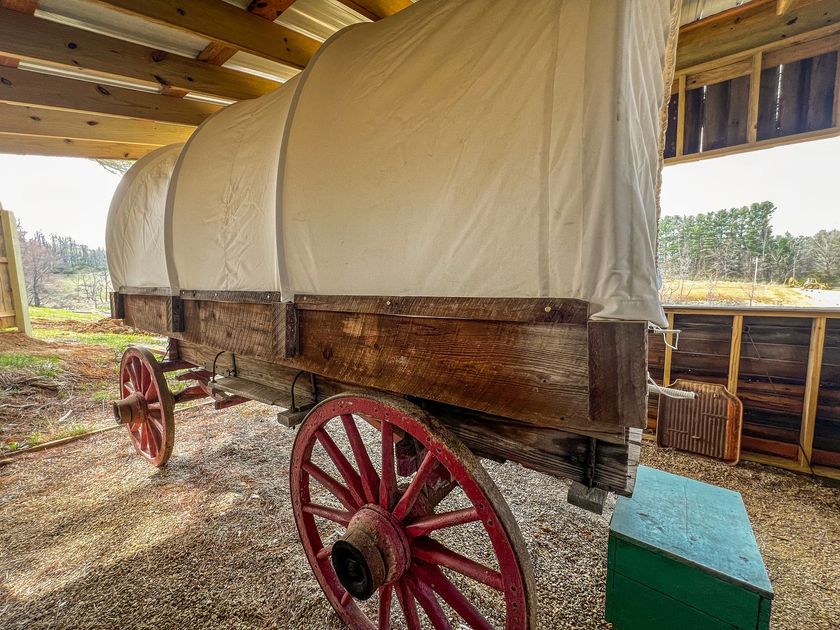 An old wooden covered wagon with red wheels is sitting in a barn.