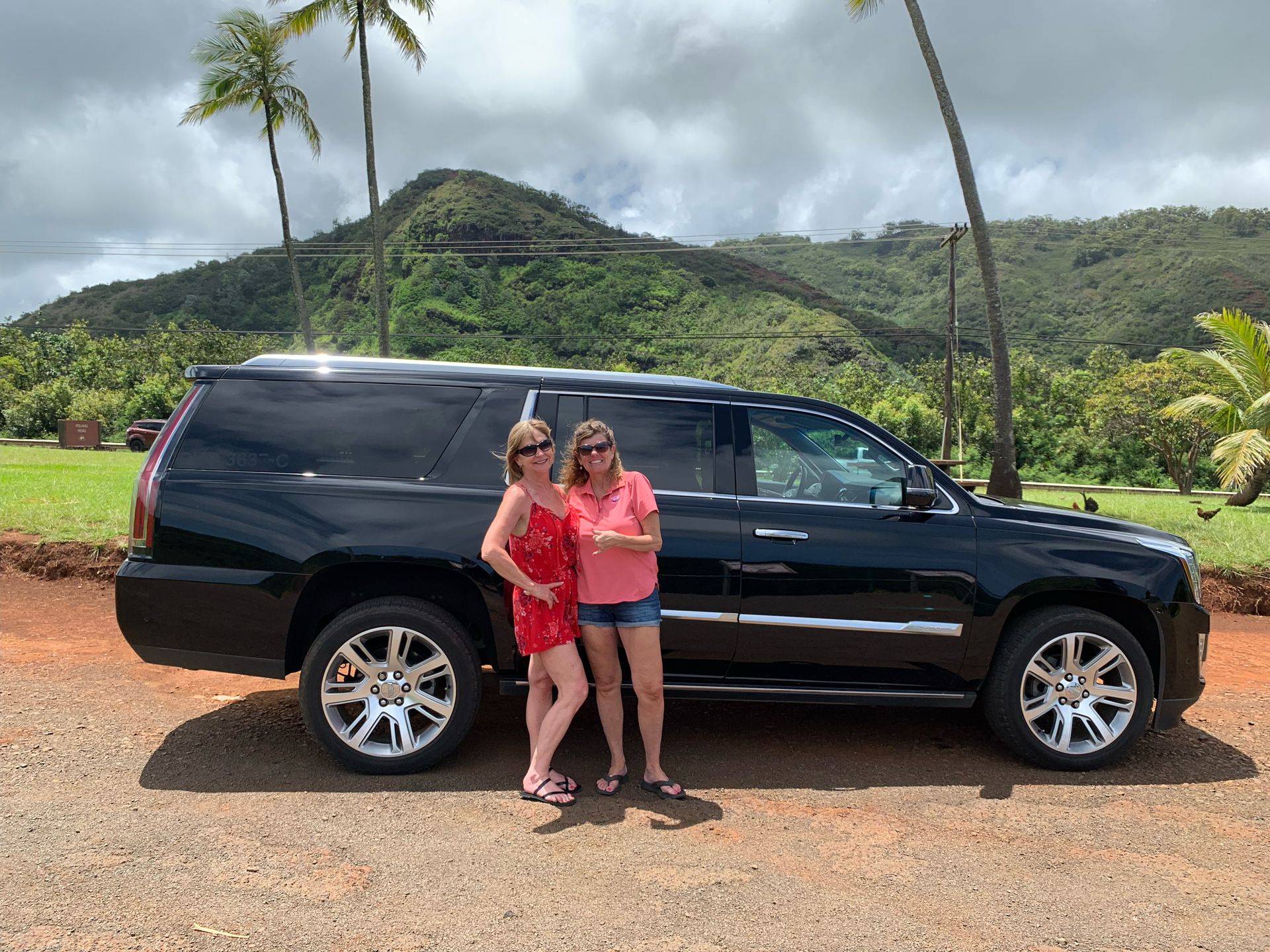 Two women stand beside a black SUV in a tropical setting with mountains and palm trees.