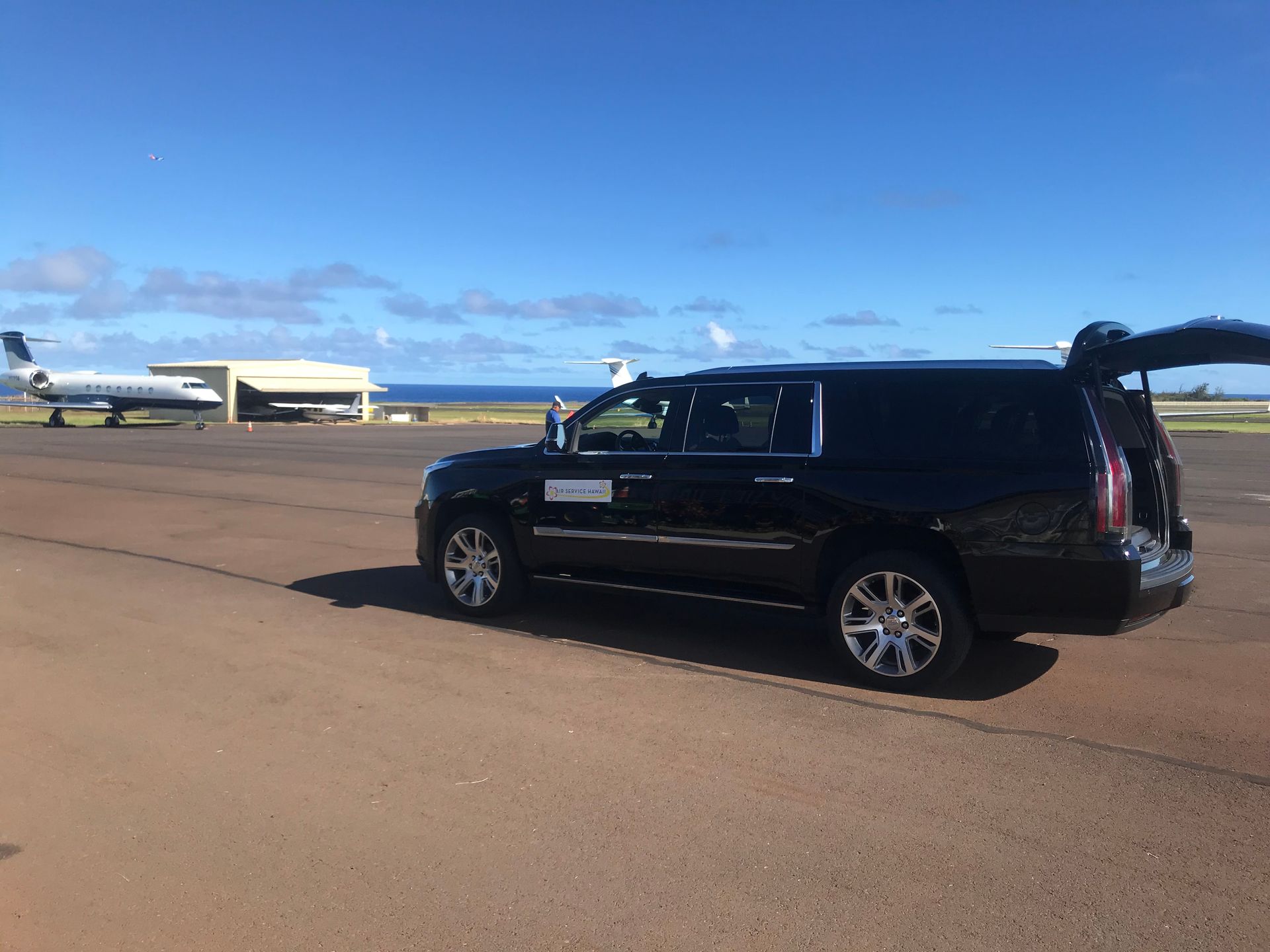 Black SUV on an airport runway, with its trunk open. A plane and hangar are in the background under a blue sky.