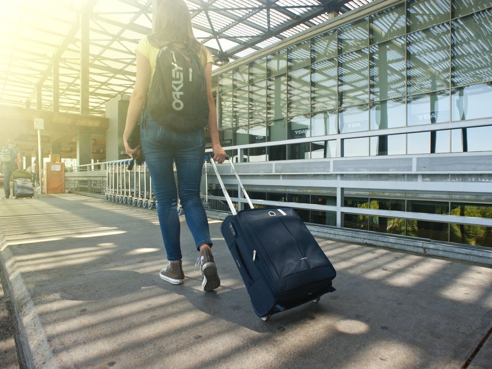Woman with backpack and wheeled suitcase walking toward airport.