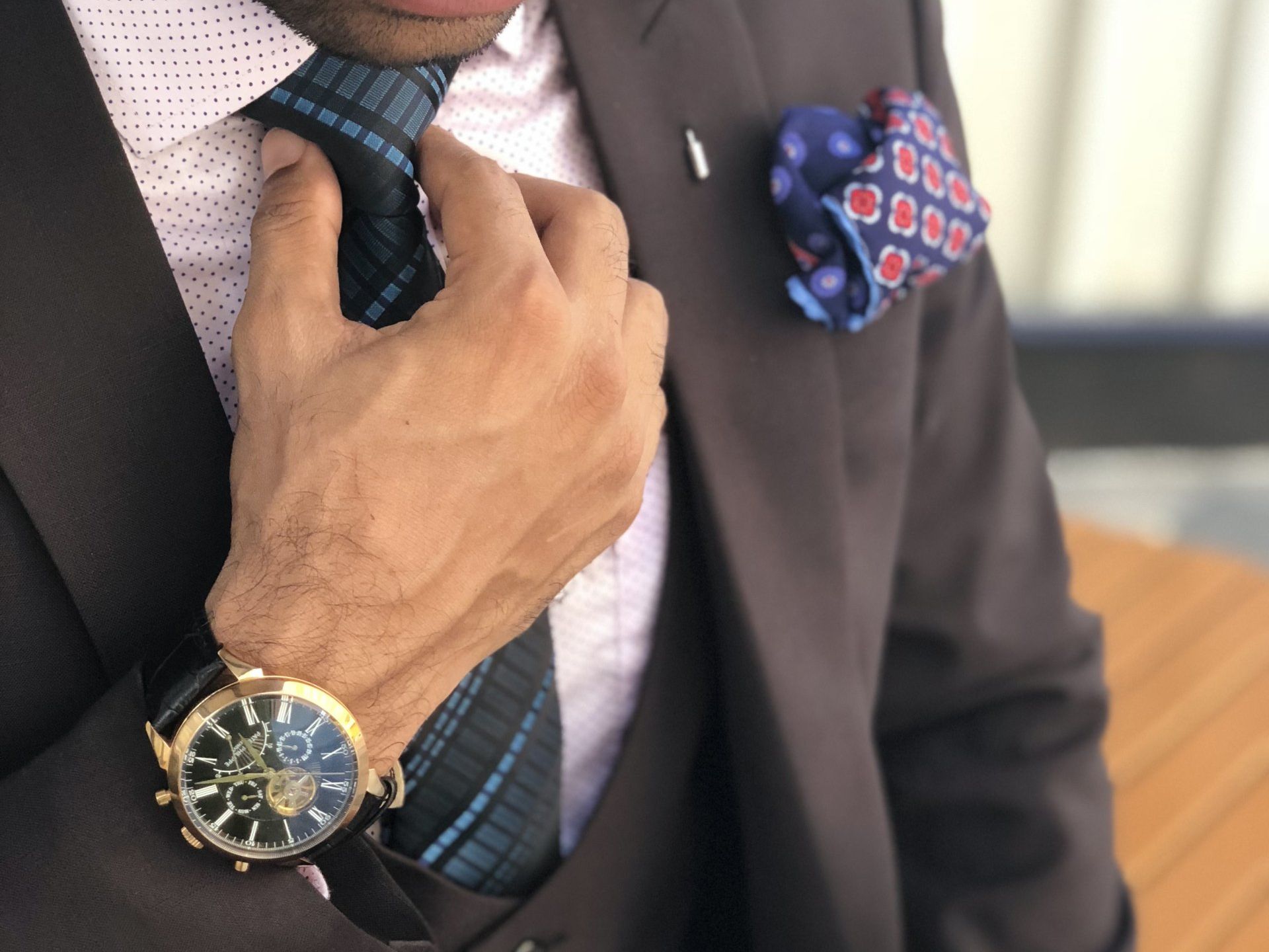 Man in brown suit adjusts tie, wearing watch, pocket square.