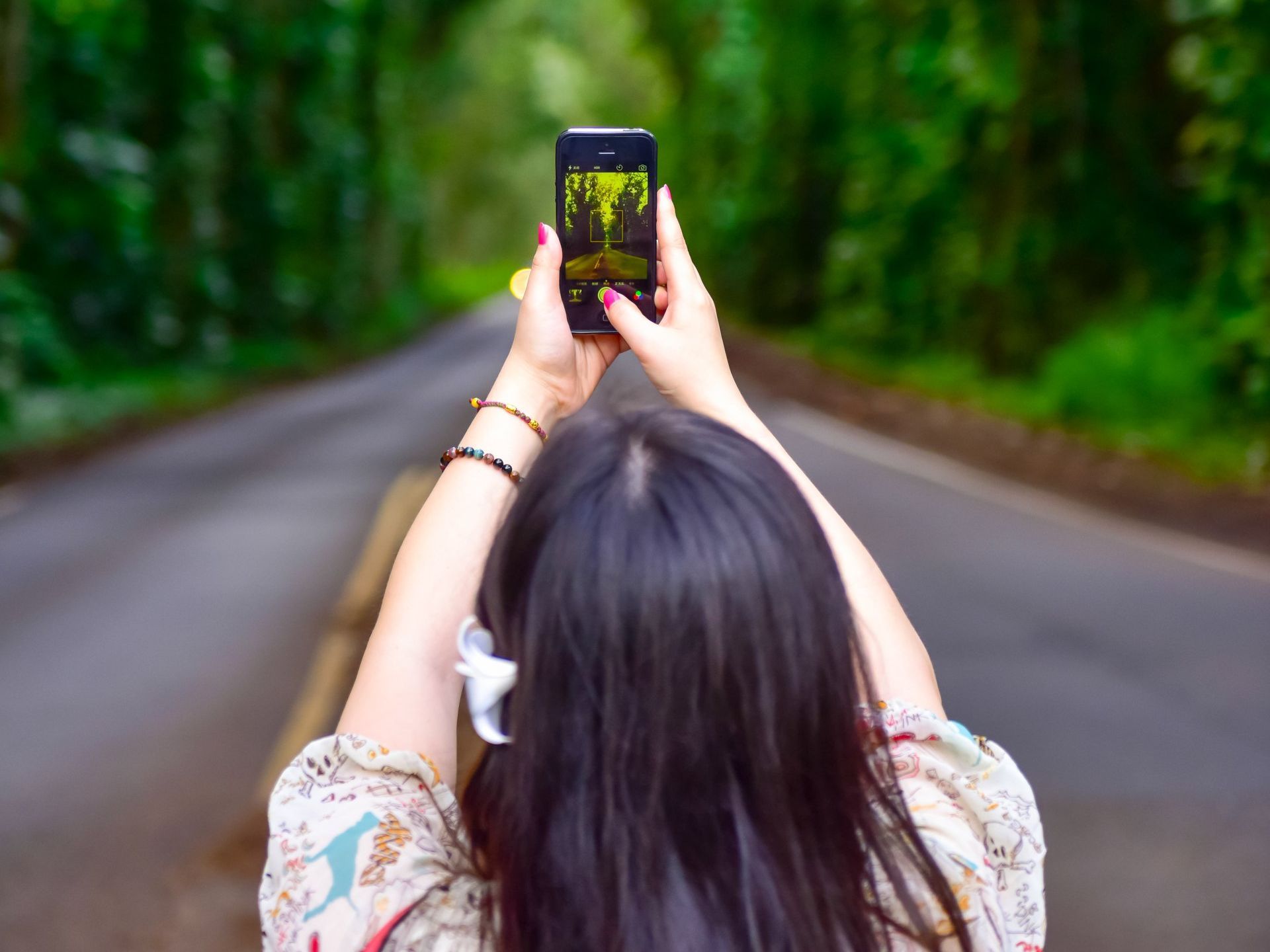 Person with dark hair takes photo of road surrounded by trees, arms outstretched, back to camera.