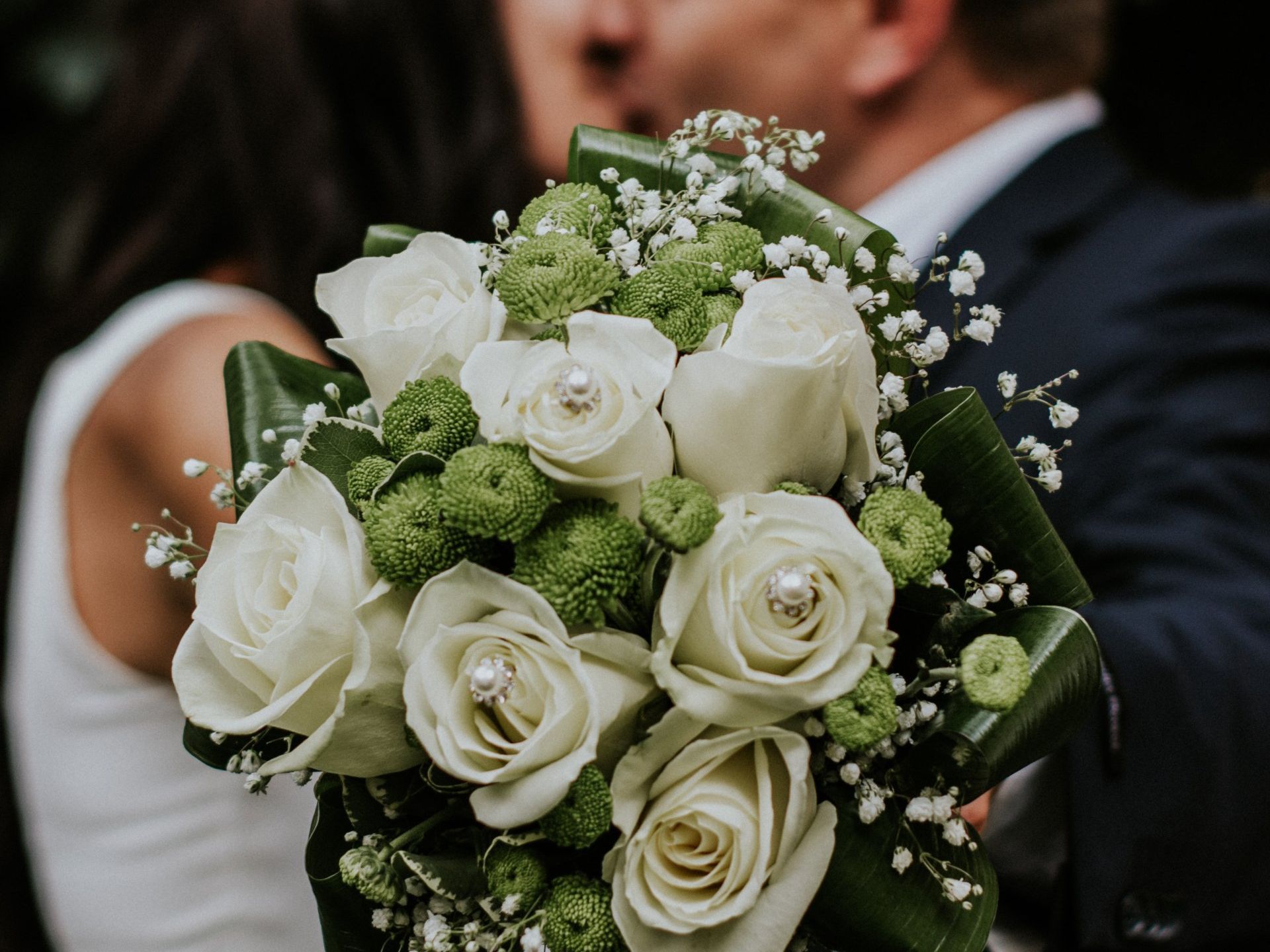 Wedding bouquet with white roses, green accents, and a couple kissing in the background.