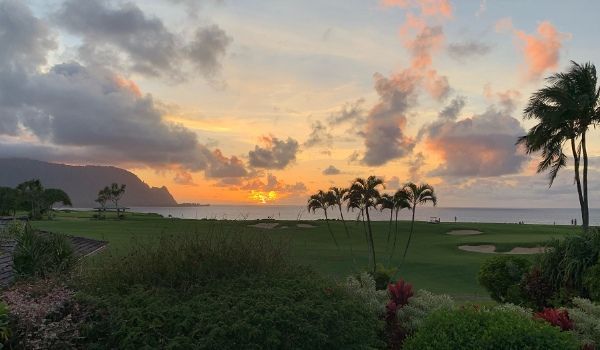 Sunset over a golf course with palm trees and ocean in the background. Cloudy sky with orange and pink hues.