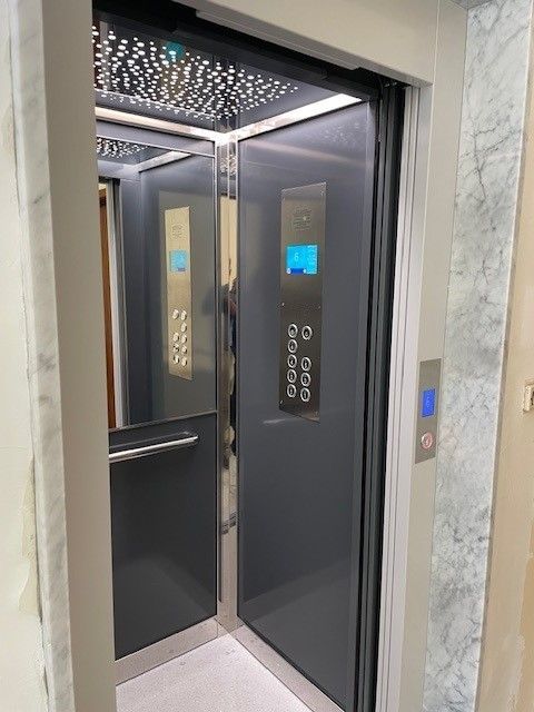 Ascensore con porte e pannello di controllo grigi, che si apre per rivelare l'interno e un corridoio. Pareti in marmo.