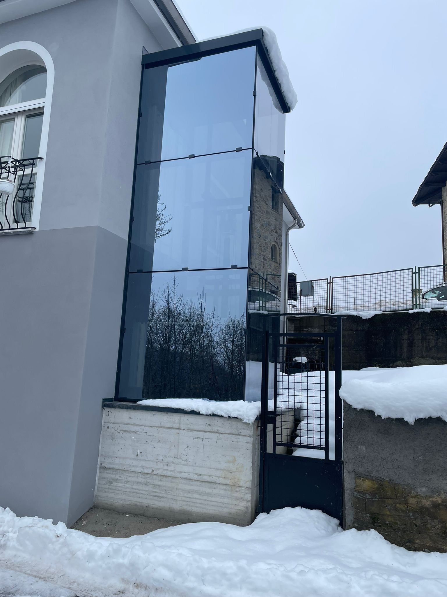 Ascensore con pareti in vetro attaccato a un edificio, accanto a un cancello nero. Neve per terra.