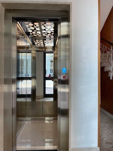 Ascensore aperto con struttura in acciaio inossidabile che riflette il corridoio, pannello decorativo sul soffitto e pulsante blu.