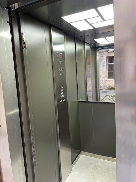 Interno di un ascensore con pareti e pulsanti in metallo grigio scuro e vista sull'esterno attraverso una porta aperta.