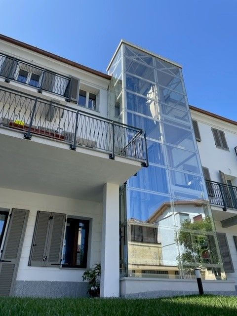 Ascensore in vetro installato all'esterno di un edificio bianco con balconi e persiane, in un'area erbosa sotto un cielo azzurro.