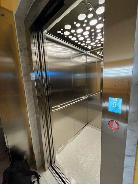 Interno dell'ascensore aperto con pareti, pavimento e soffitto in metallo lucido con luci a bolla. La pulsantiera mostra il numero 5.
