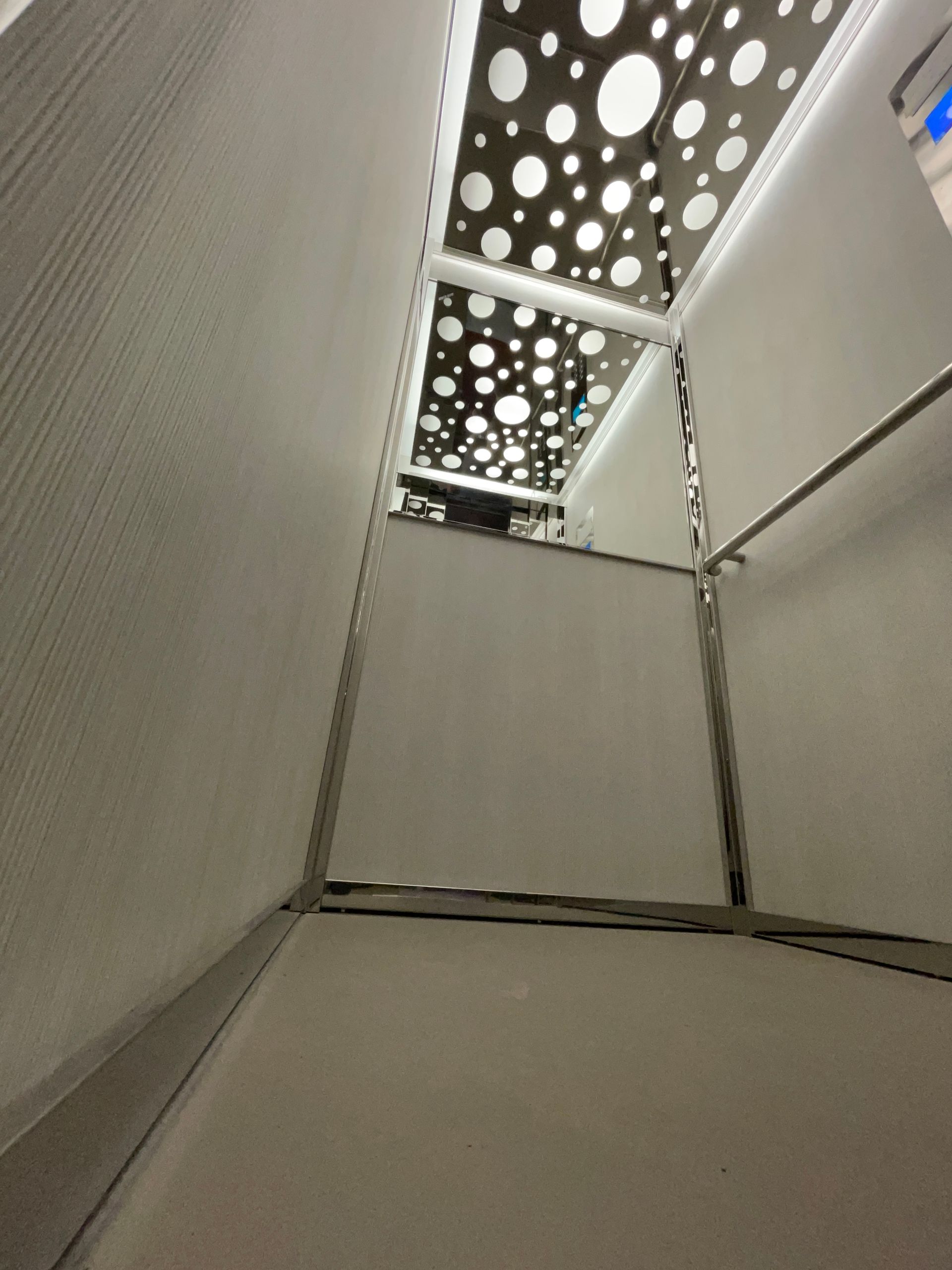 Vista dall'alto all'interno di un moderno ascensore. Pareti bianche, struttura in acciaio inossidabile, soffitto perforato con cerchi.