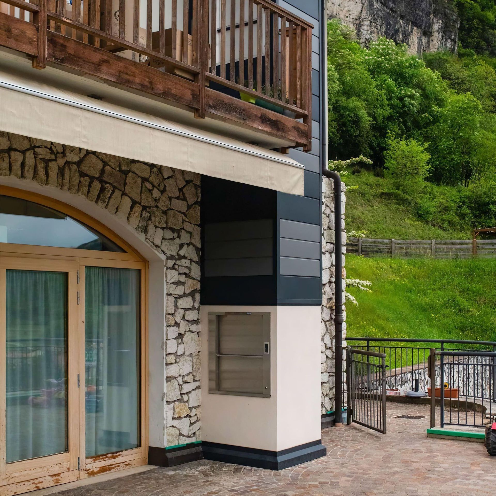 Esterno dell'edificio con muro in pietra, balcone in legno e pendio erboso.