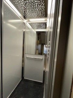 All'interno di un moderno ascensore: pareti bianche, soffitto con ritagli a forma di stella, corrimano in metallo, porta aperta.