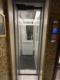 Interno di un ascensore con una persona riflessa nello specchio. Rifiniture in metallo lucido, pareti bianche e tappetino nero.