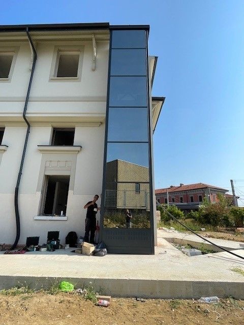 Ascensore esterno sul lato di un edificio, vetro a specchio. Una persona ci sta lavorando. Cielo azzurro e limpido.