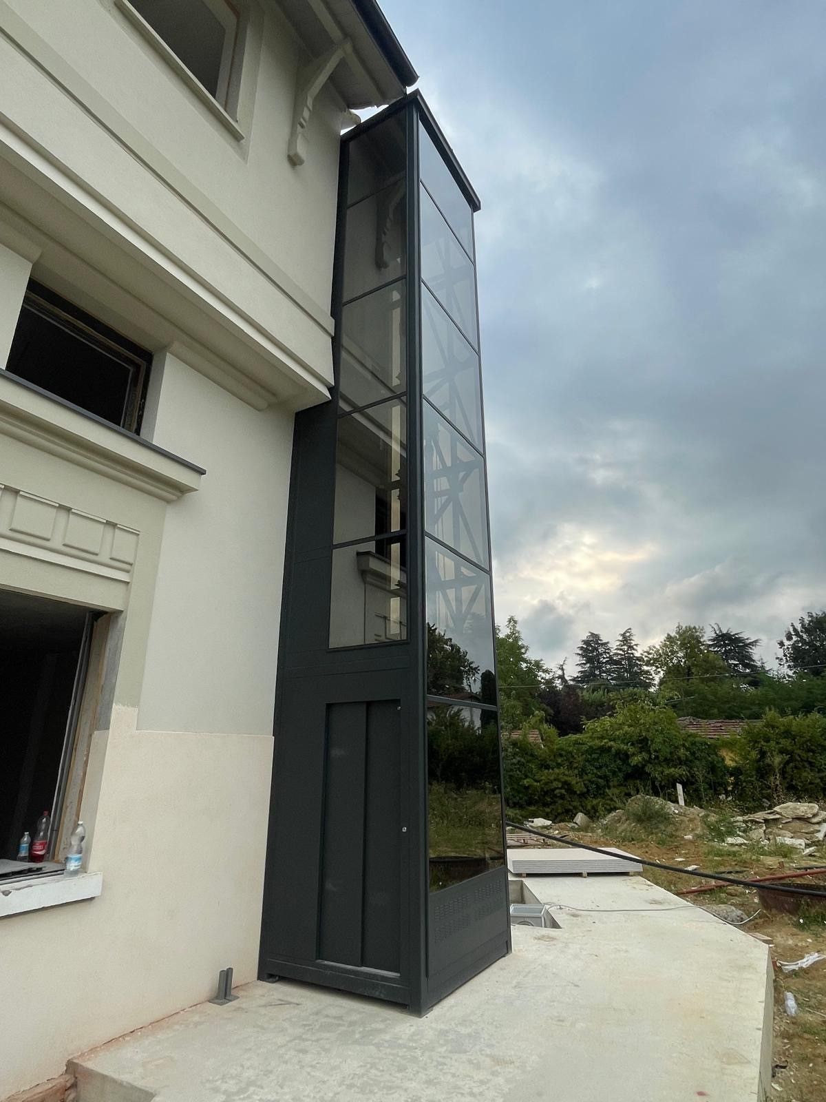 Ascensore residenziale esterno in vetro collegato a un edificio di colore chiaro. L'ascensore è grigio scuro/nero con base in cemento. Cielo nuvoloso.