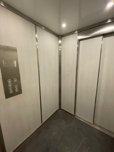 Interno di un ascensore con pareti bianche, finiture in acciaio inossidabile e pannello di controllo sulla sinistra.