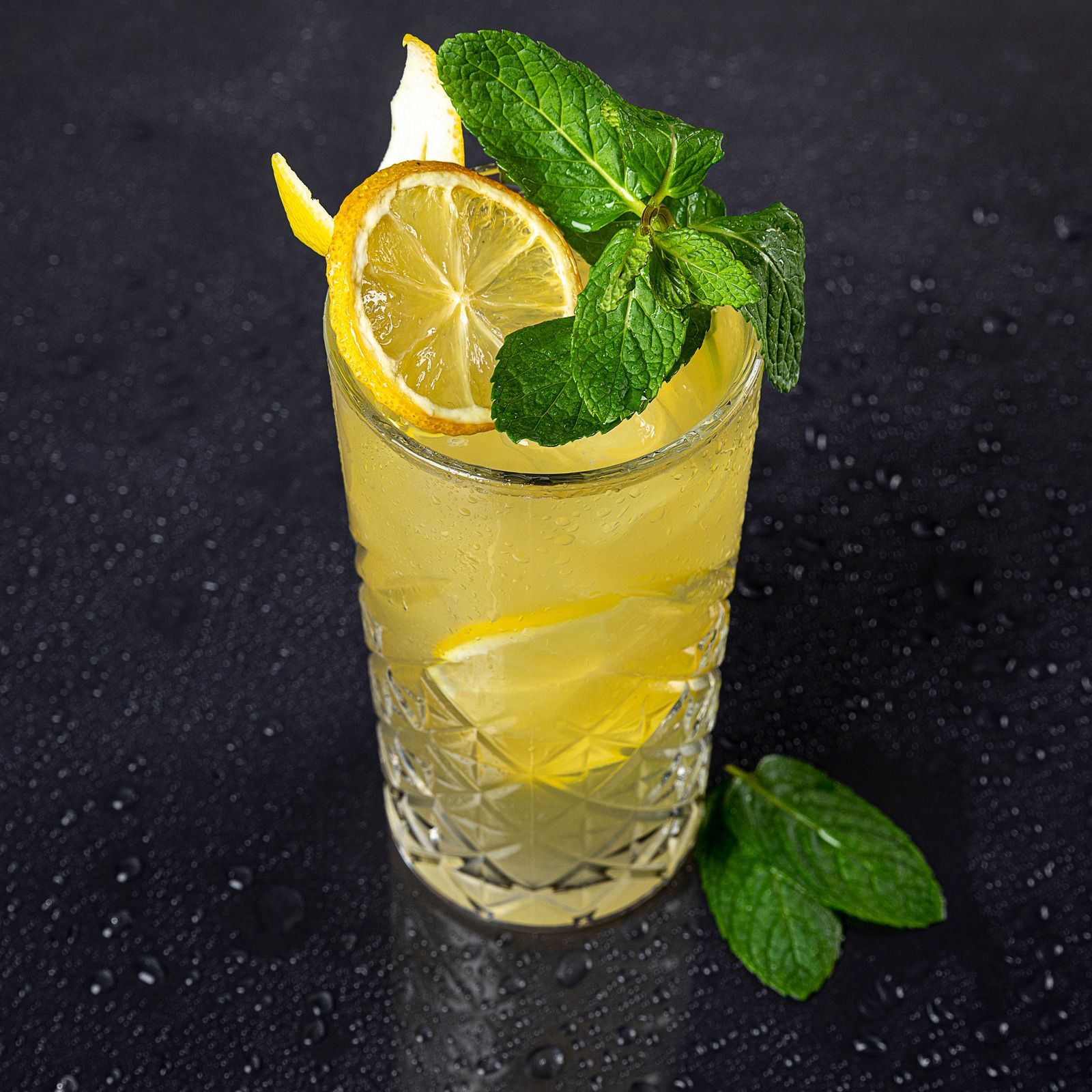 Yellow cocktail in a textured glass, garnished with lemon slice and mint sprig, on a wet, dark surface.