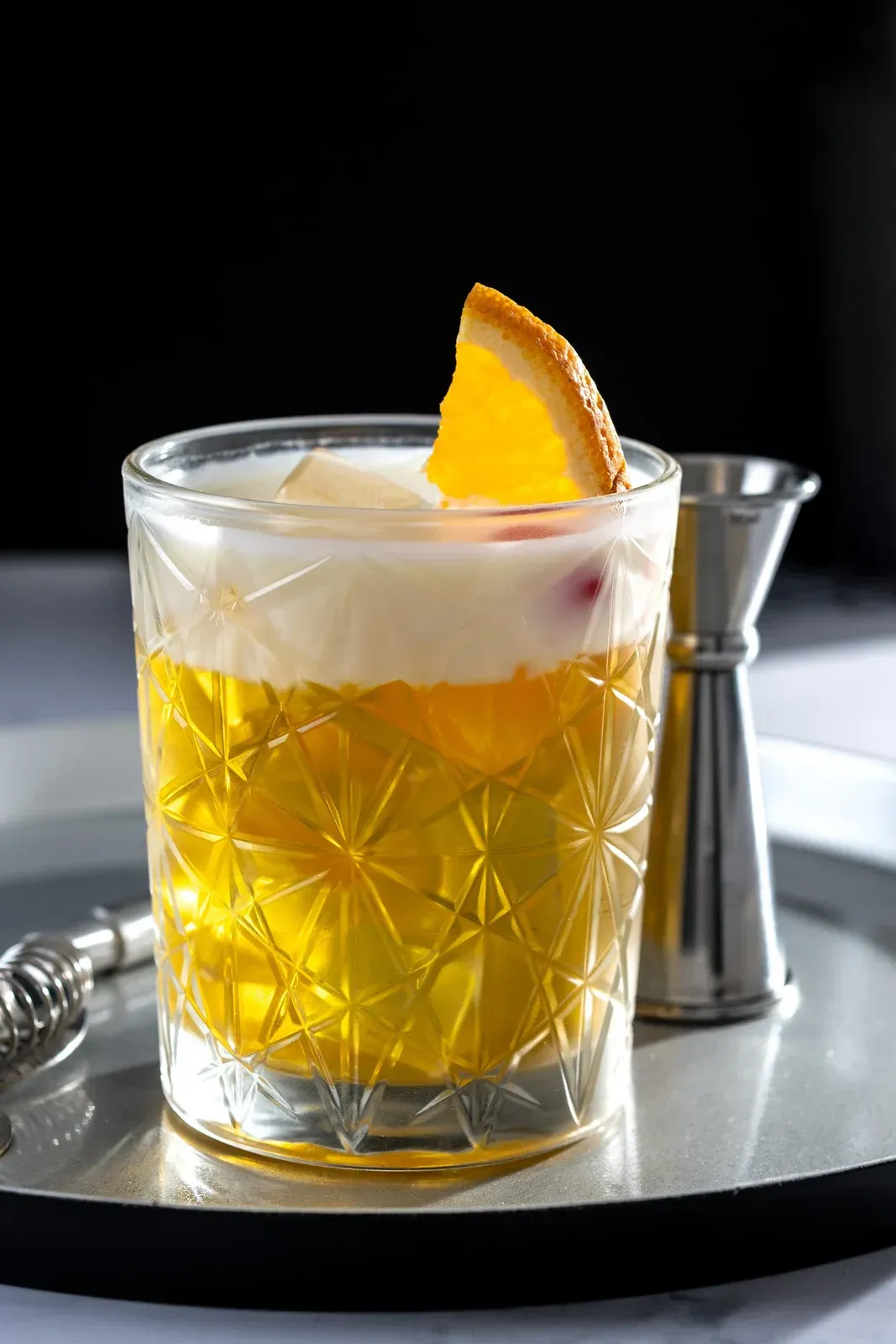 Cocktail in a cut-glass tumbler with foamy top and orange slice garnish, on a silver tray with a jigger.