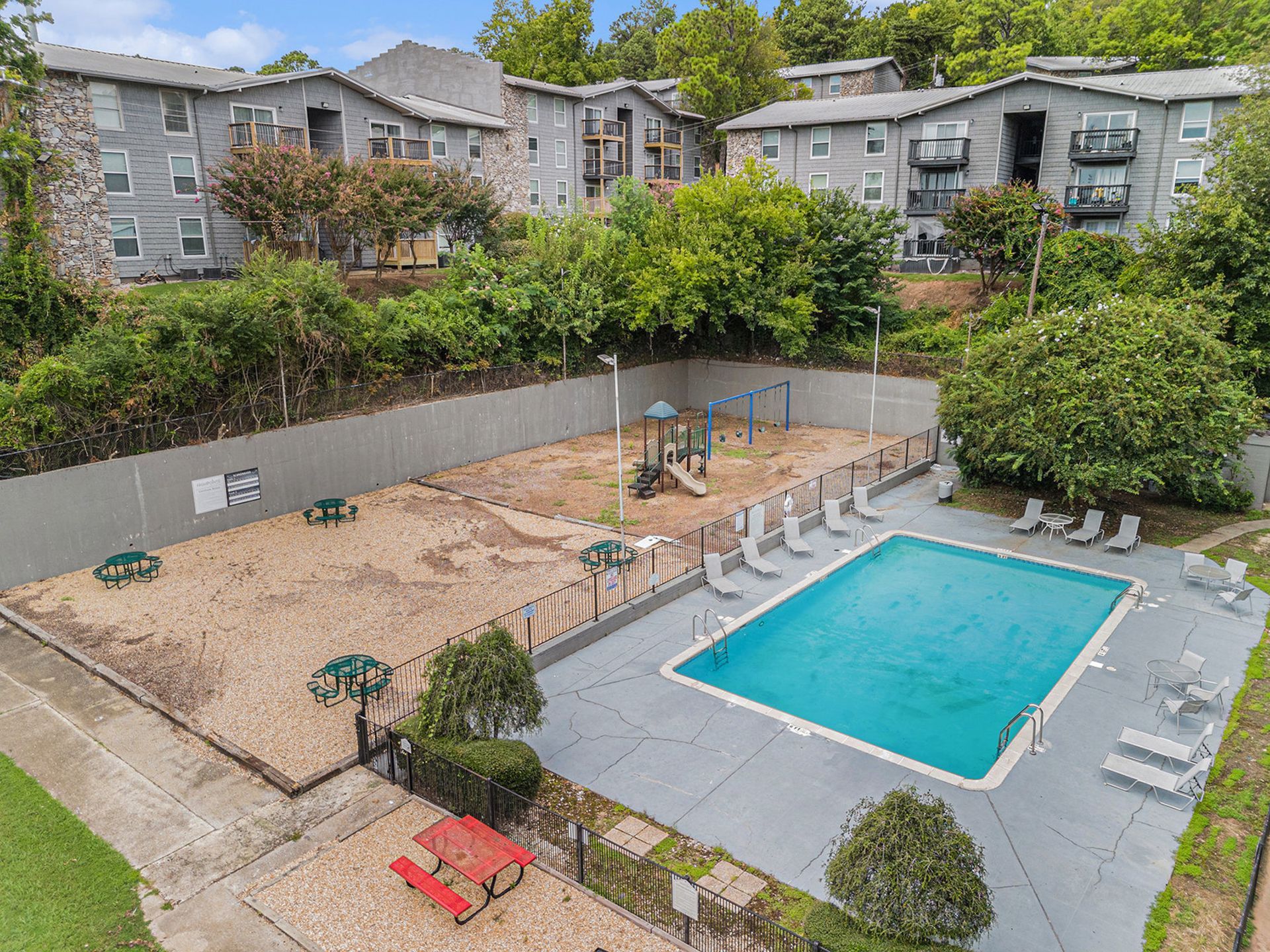 Aerial view of a rectangular swimming pool next to a fenced playground area with wood chips and apartment buildings.