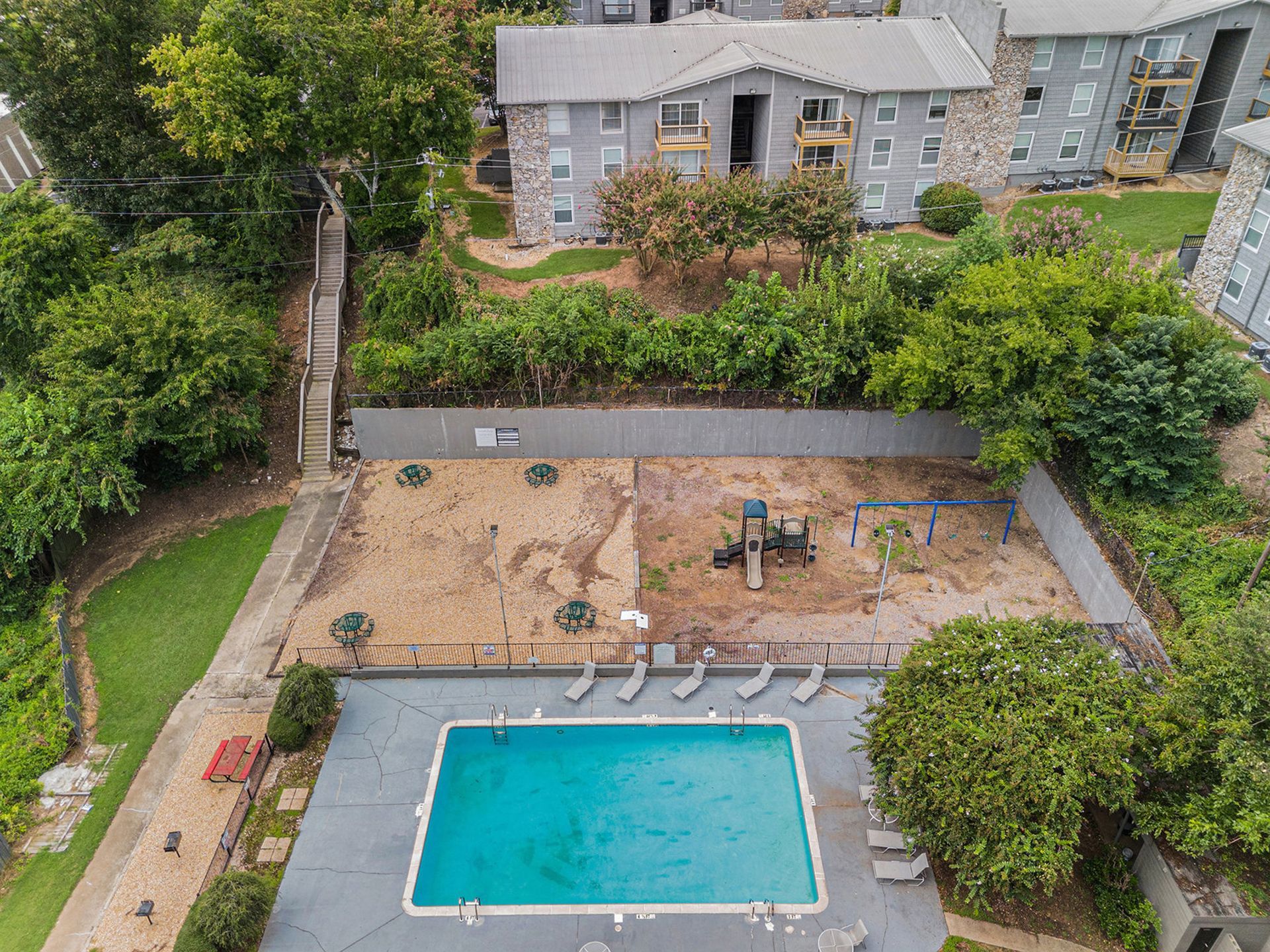 High-angle view of a rectangular swimming pool next to a playground area with mulch, trees, and apartment buildings.