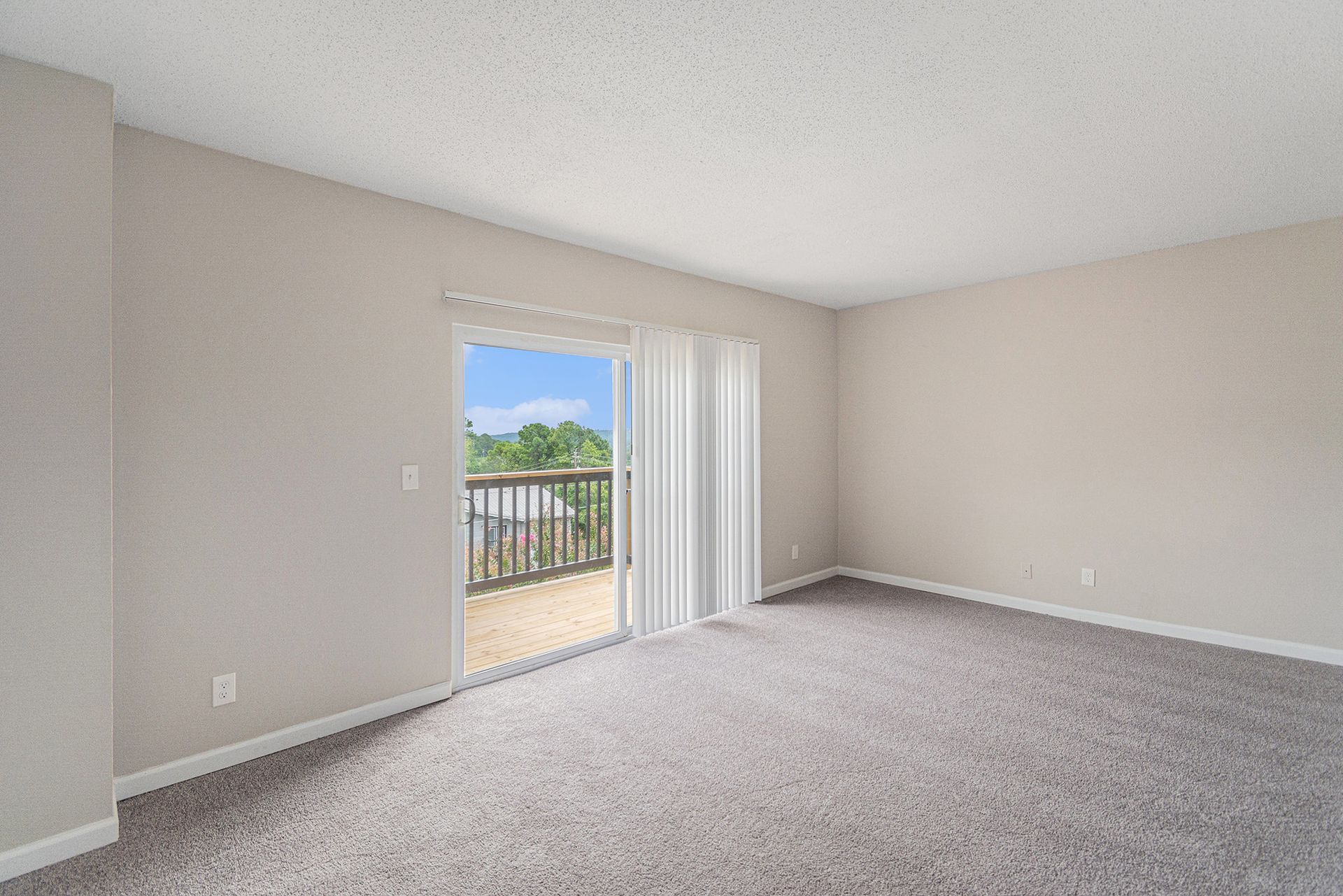 An empty room with neutral beige walls, gray carpeting, and a sliding glass door opening to a balcony.