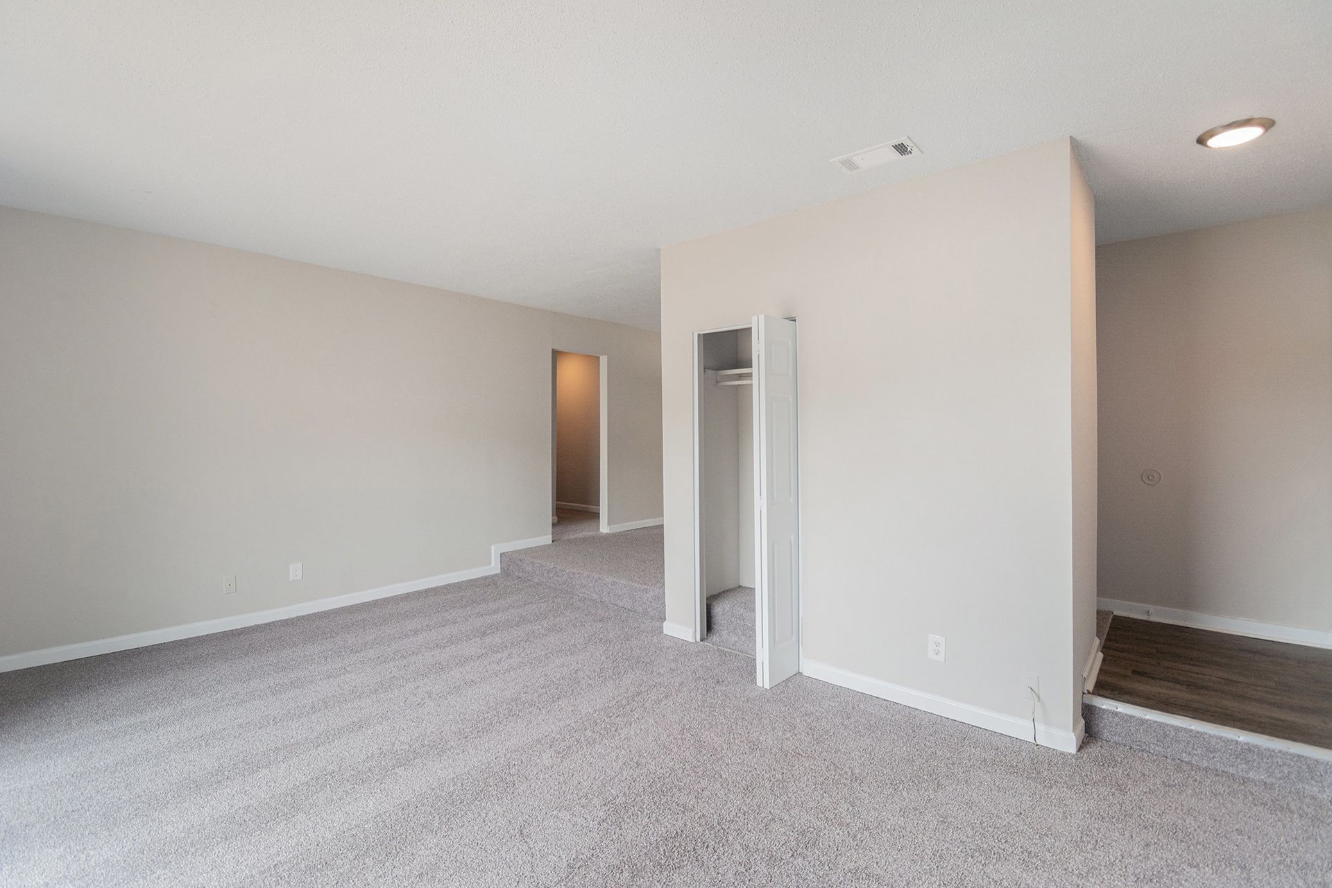 Empty carpeted room with light gray walls, a small recessed closet, and an adjacent area with hardwood flooring.
