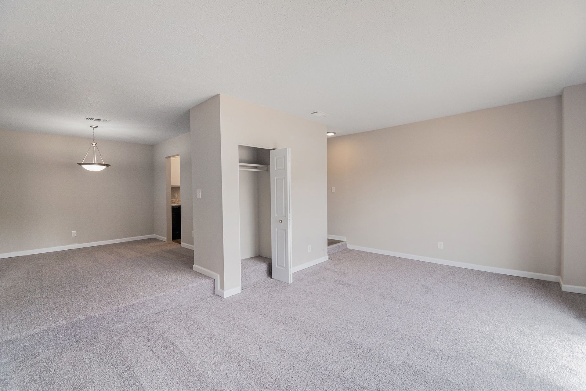 An empty room with beige walls and gray carpet, featuring an open doorway to a closet and an adjacent dining area.