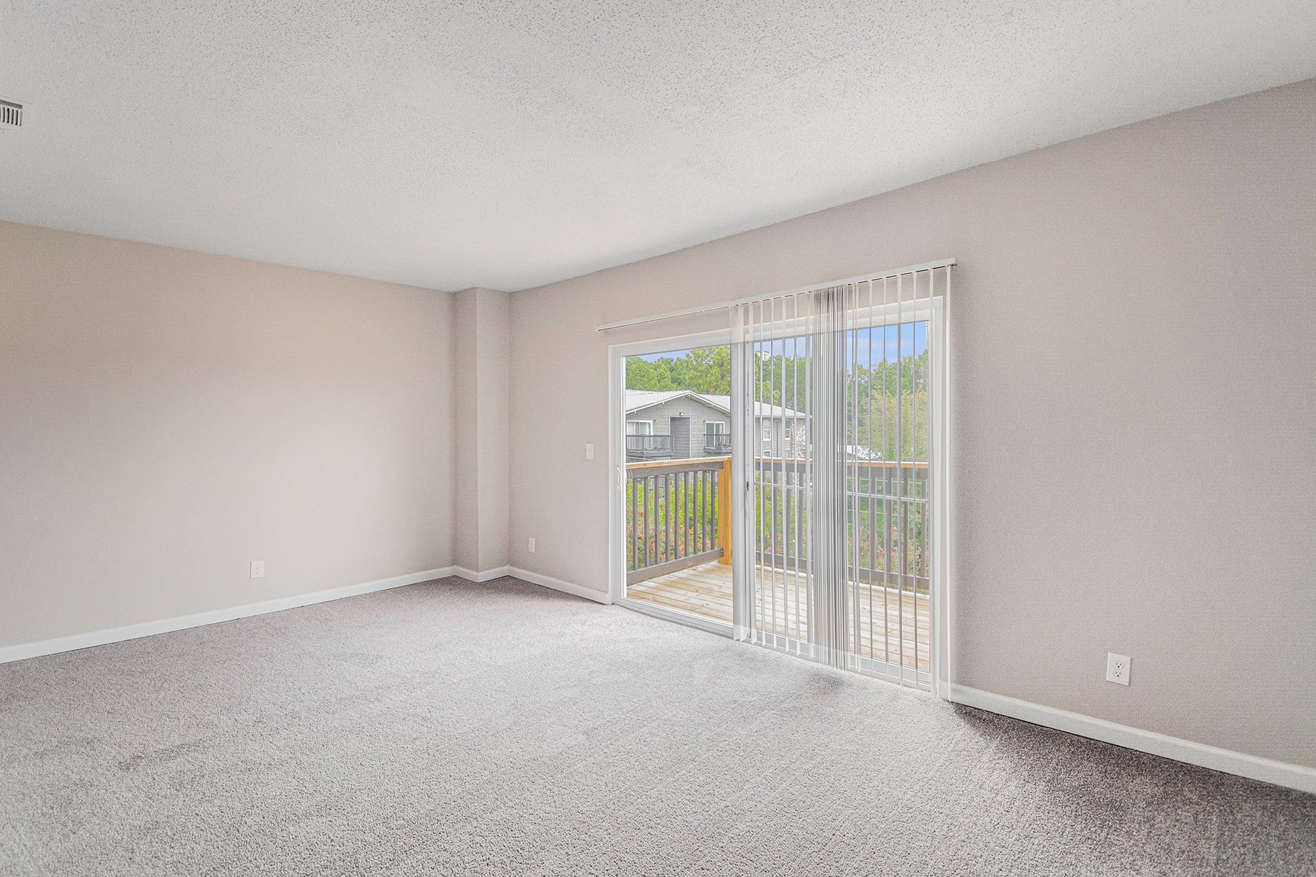 An empty, carpeted room with beige walls and a sliding glass door leading to a wooden balcony.